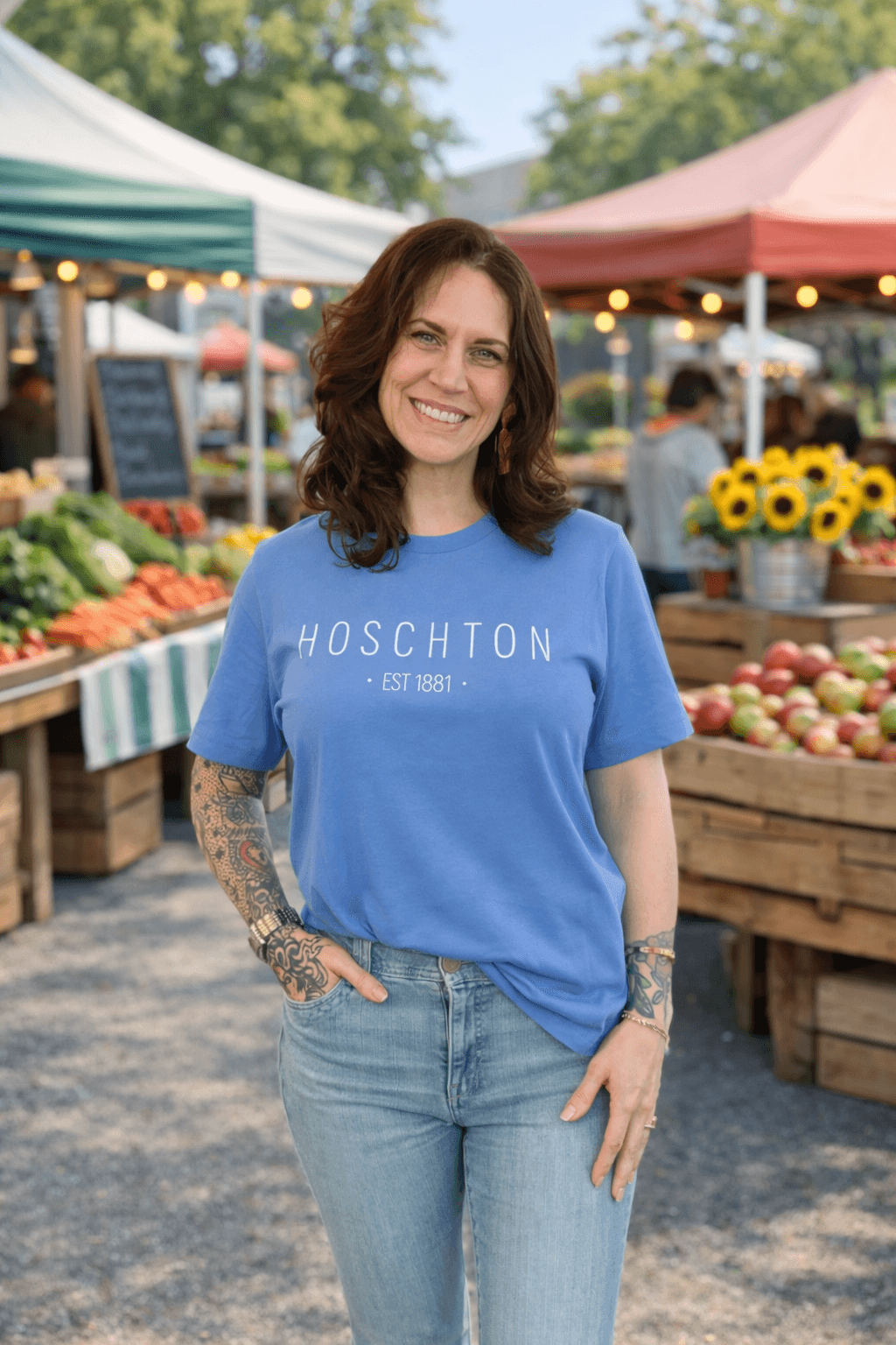 Smiling woman with wavy brown hair wearing a blue T-shirt and jeans at an outdoor market with produce and flowers.