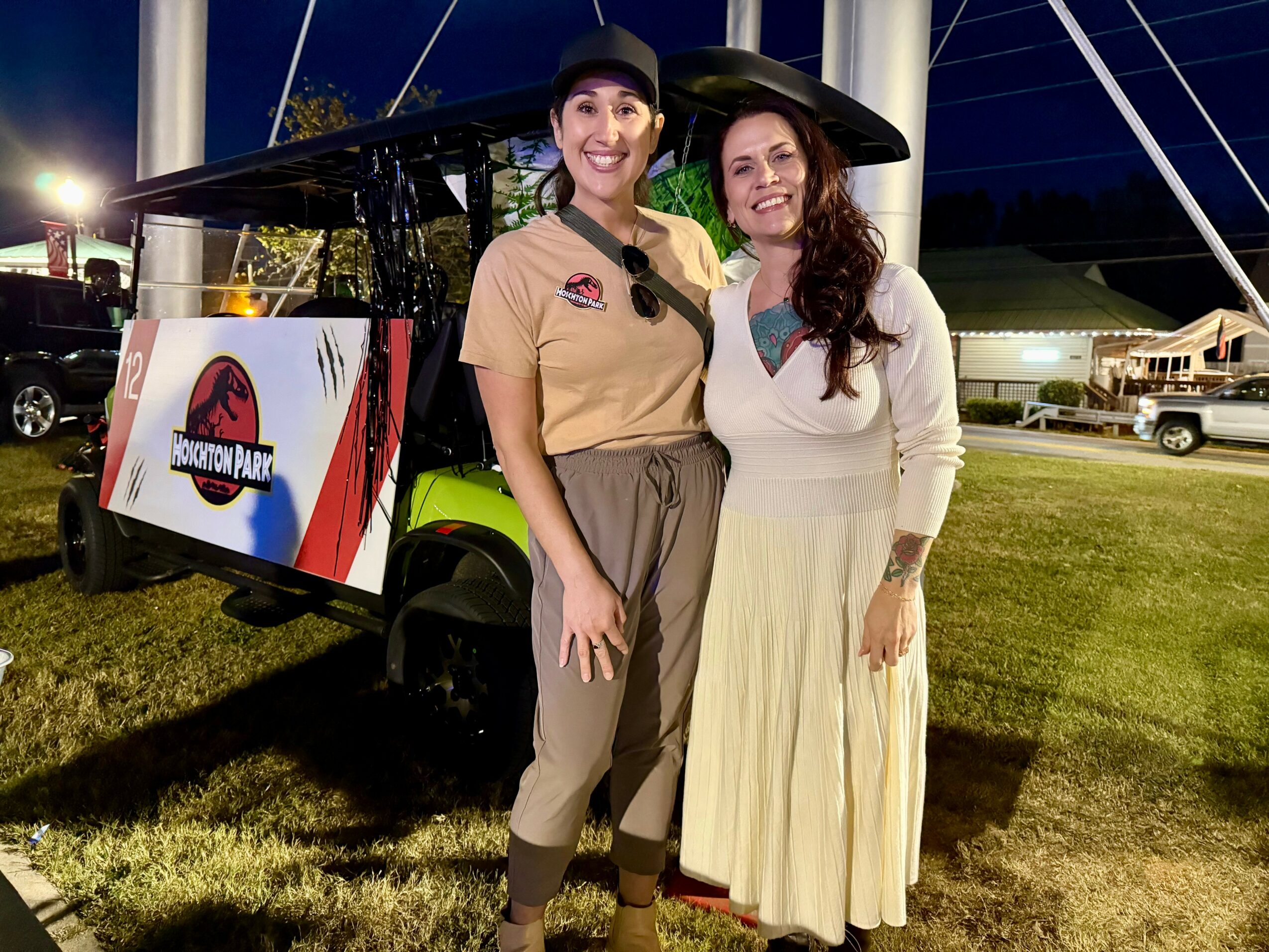 Two women stand outdoors at night, smiling, with a golf cart and a sign in the background.
