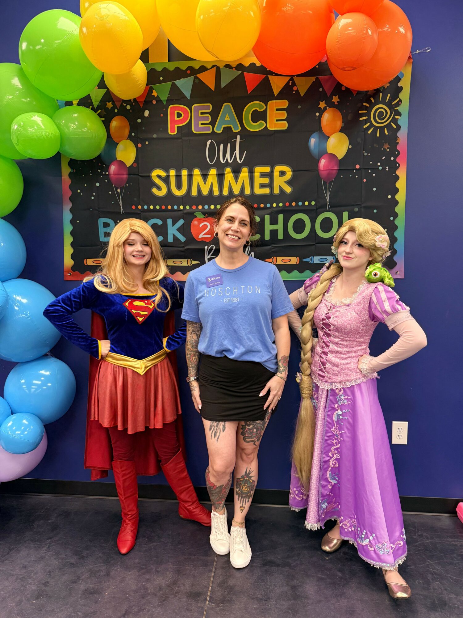 Three women in colorful costumes pose at a summer party with balloons and a decorated backdrop.
