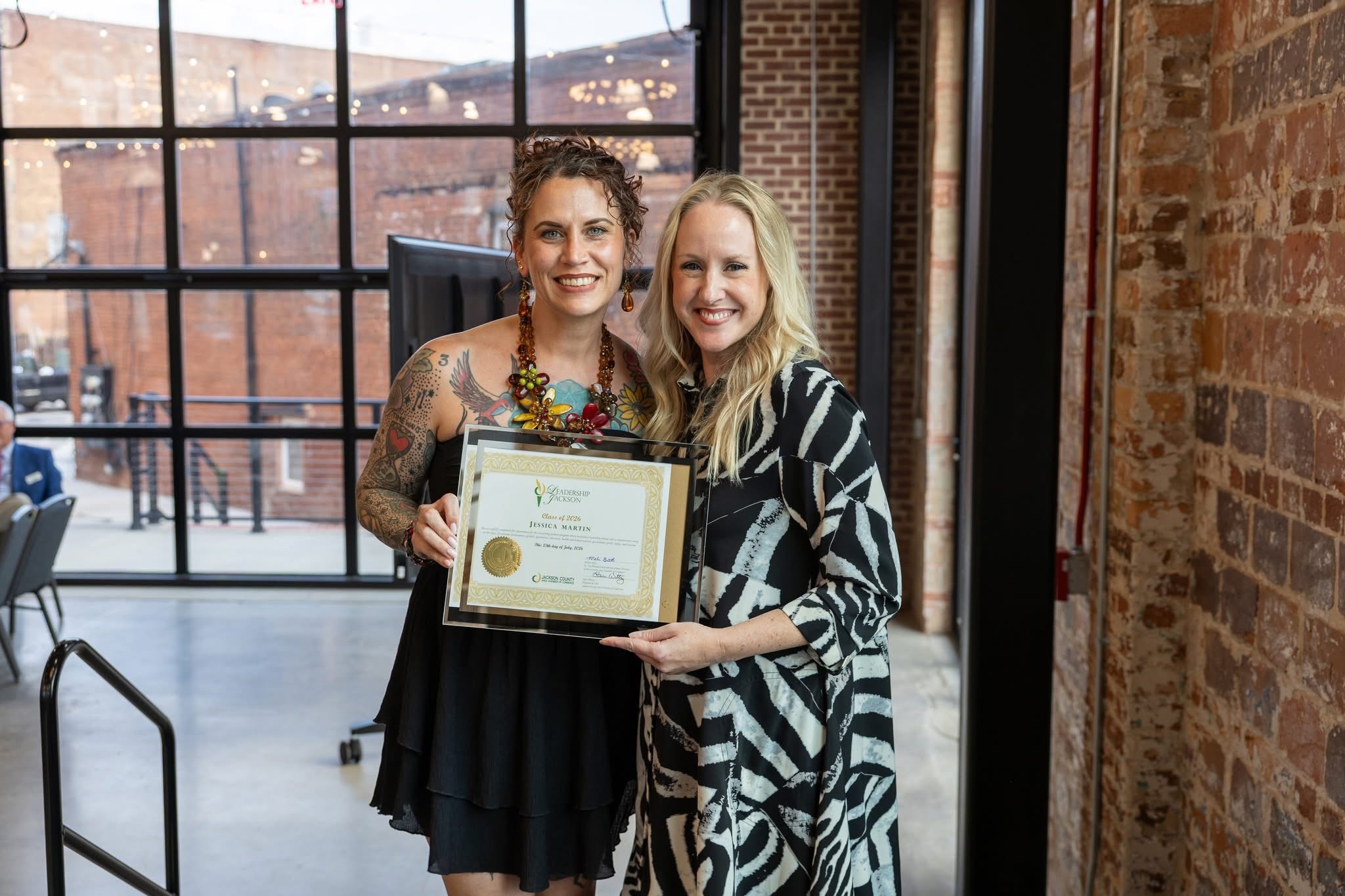 Two women smiling, one holding a certificate, standing inside a brick-walled room with large windows behind them.