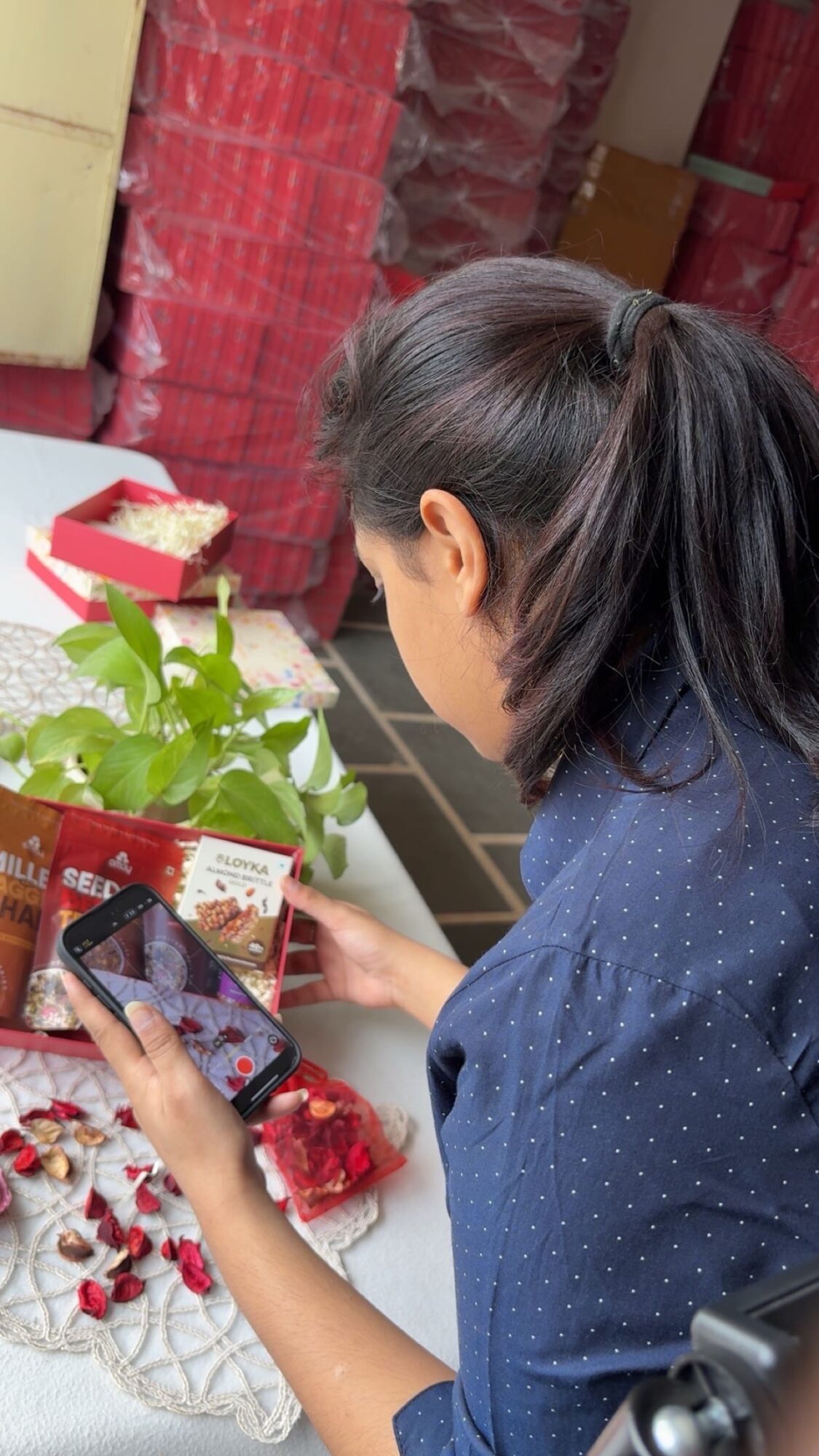Woman with dark hair in a ponytail looks at her phone, surrounded by plants and decorative items, indoors.