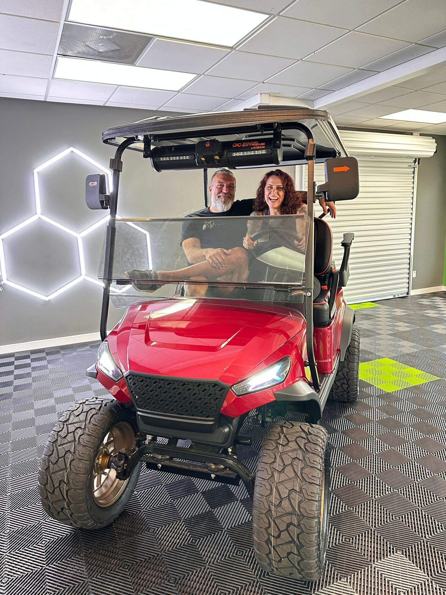 People riding a red golf cart indoors, with a person in front and others behind, on a patterned floor.