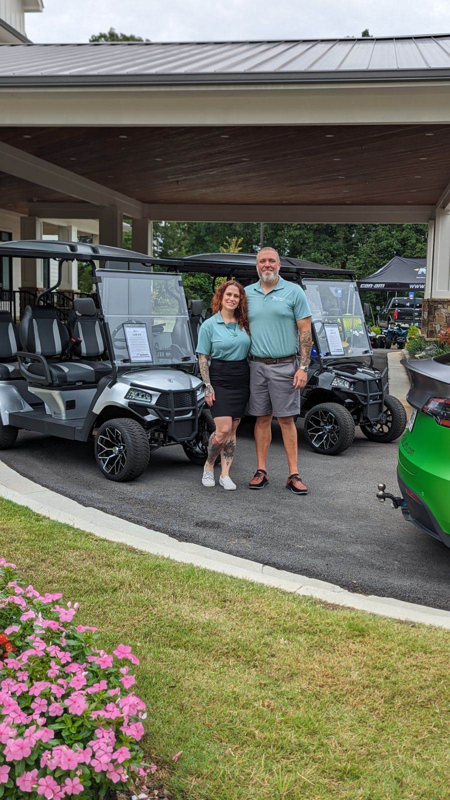 Two people stand between two golf carts under a covered area, with a green vehicle partially visible on the right.