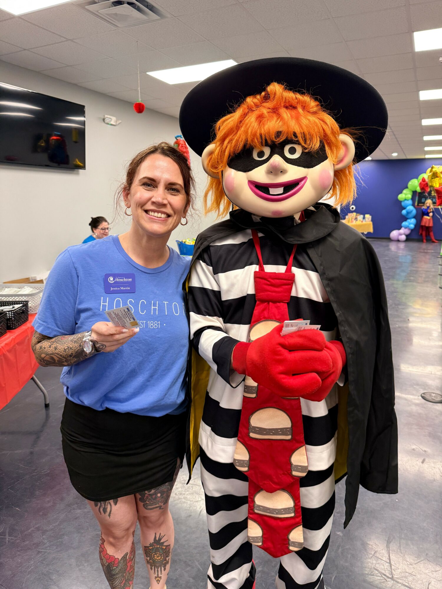 Person with orange hair and makeup dressed as a clown with a black hat, striped outfit, and red gloves, standing next to a woman in a blue shirt with tattoos, in an indoor setting.
