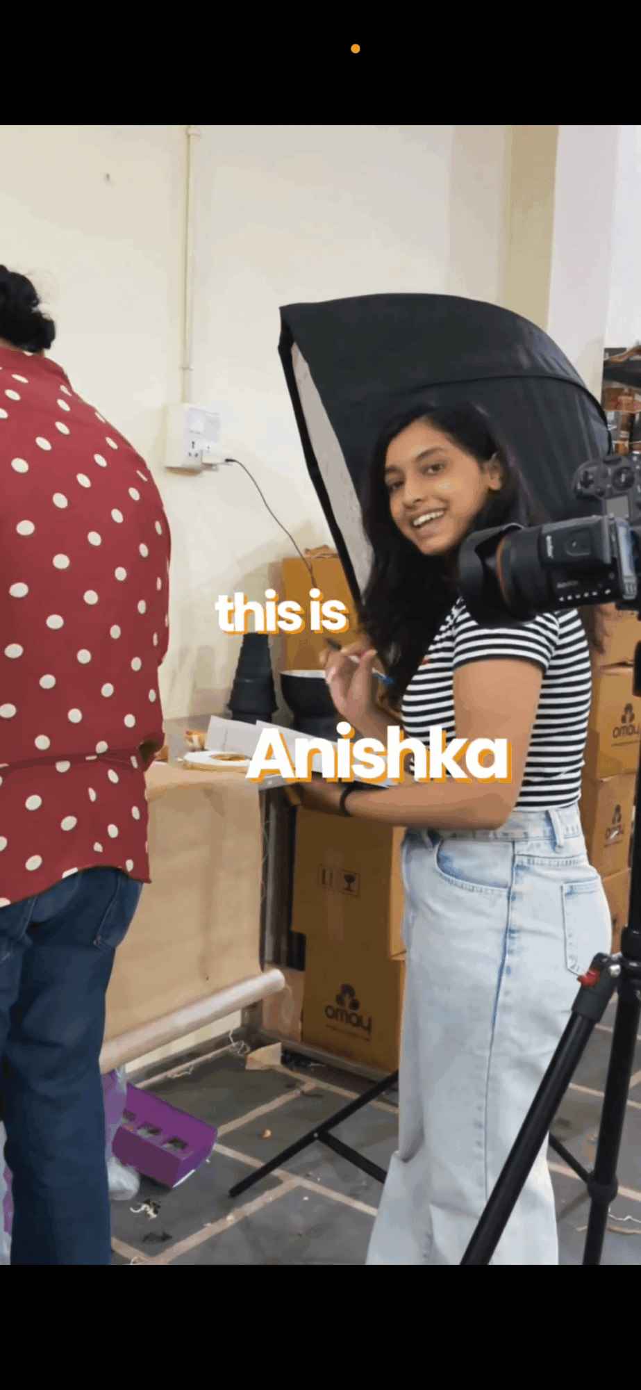 Young woman smiling at camera, holding a professional camera, in a room with boxes and a person in a red polka dot shirt.
