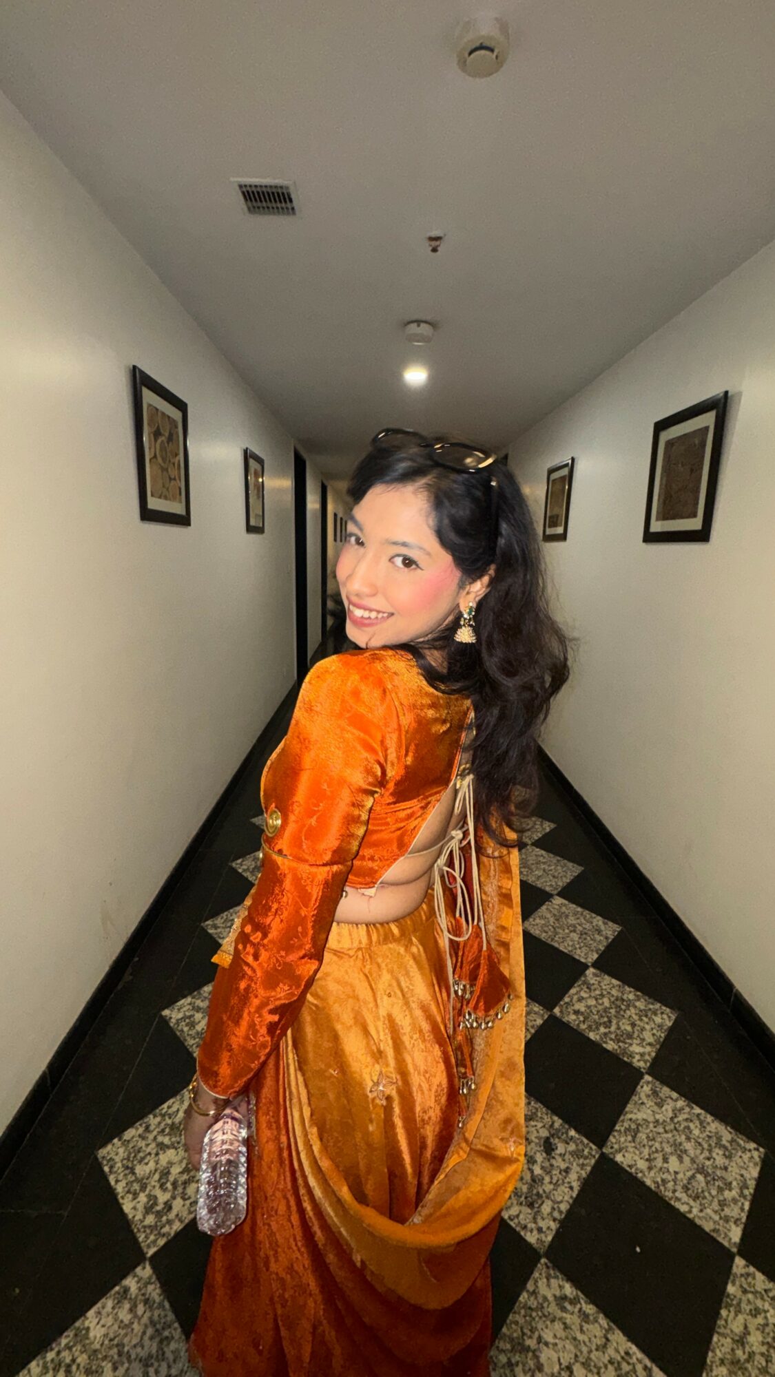Woman in orange traditional dress standing in a hallway with framed pictures, smiling at the camera.