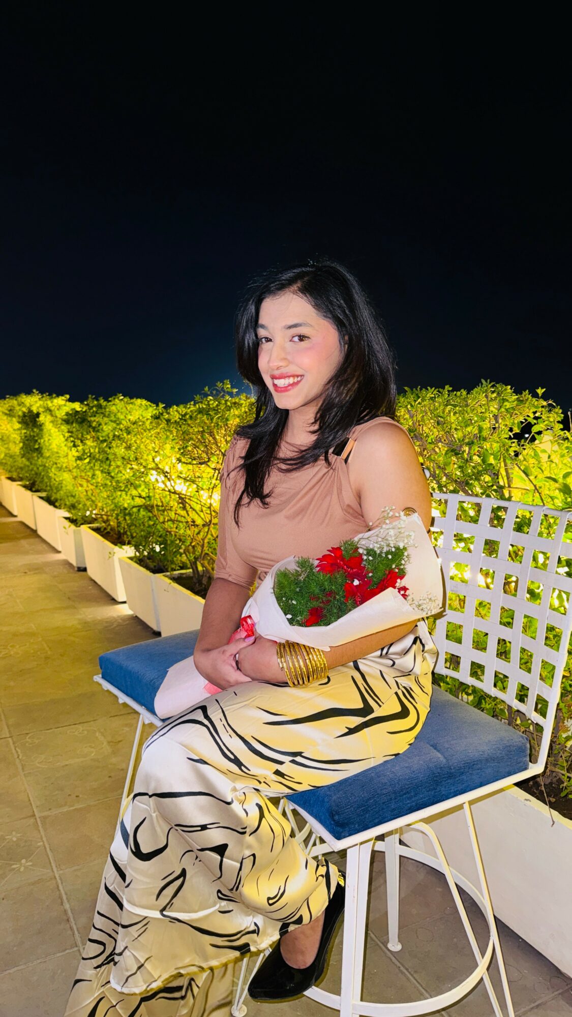 Woman sitting on a bench holding a bouquet of red and white flowers, smiling, with greenery in the background at night.