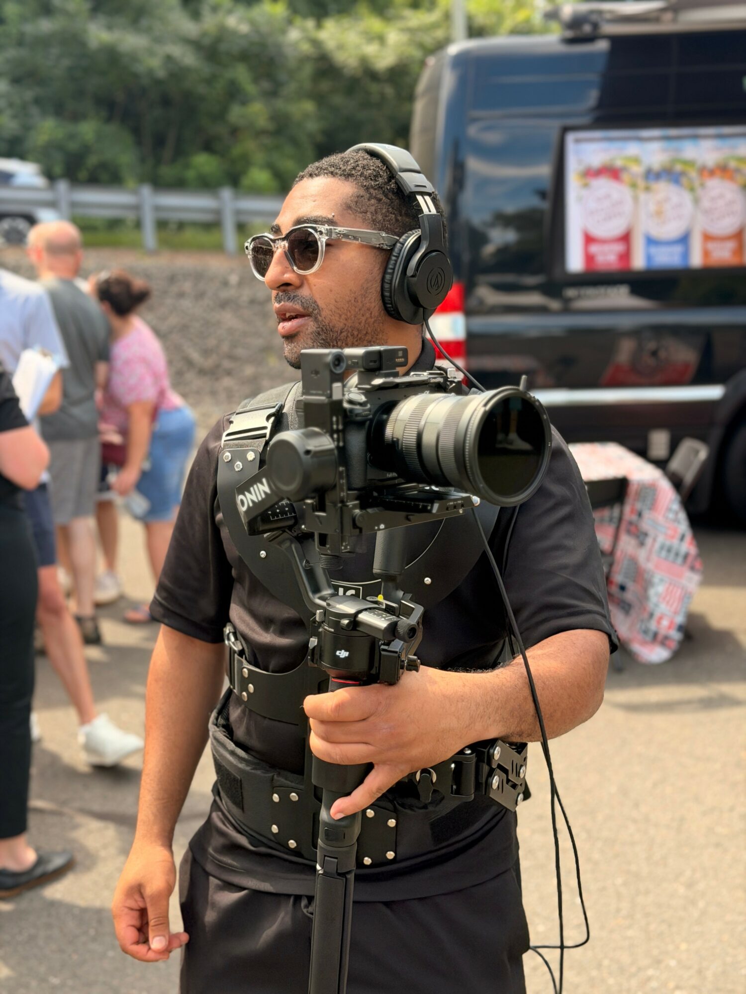 Man with sunglasses and headphones holding a professional video camera outdoors, with people and vehicles in the background.
