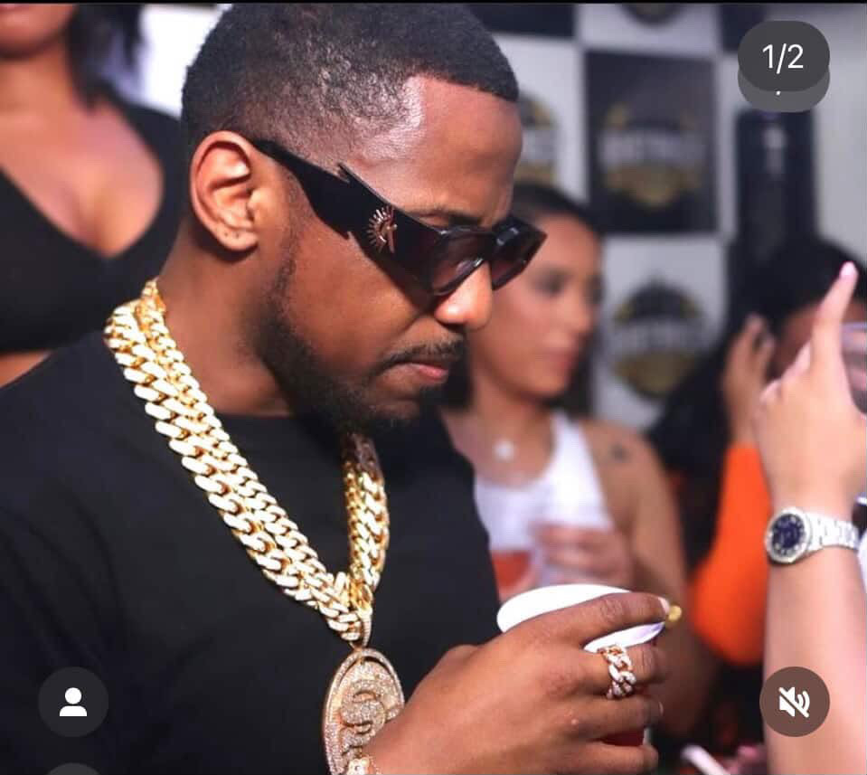 Man with sunglasses and gold chains looking at his phone in a social setting.