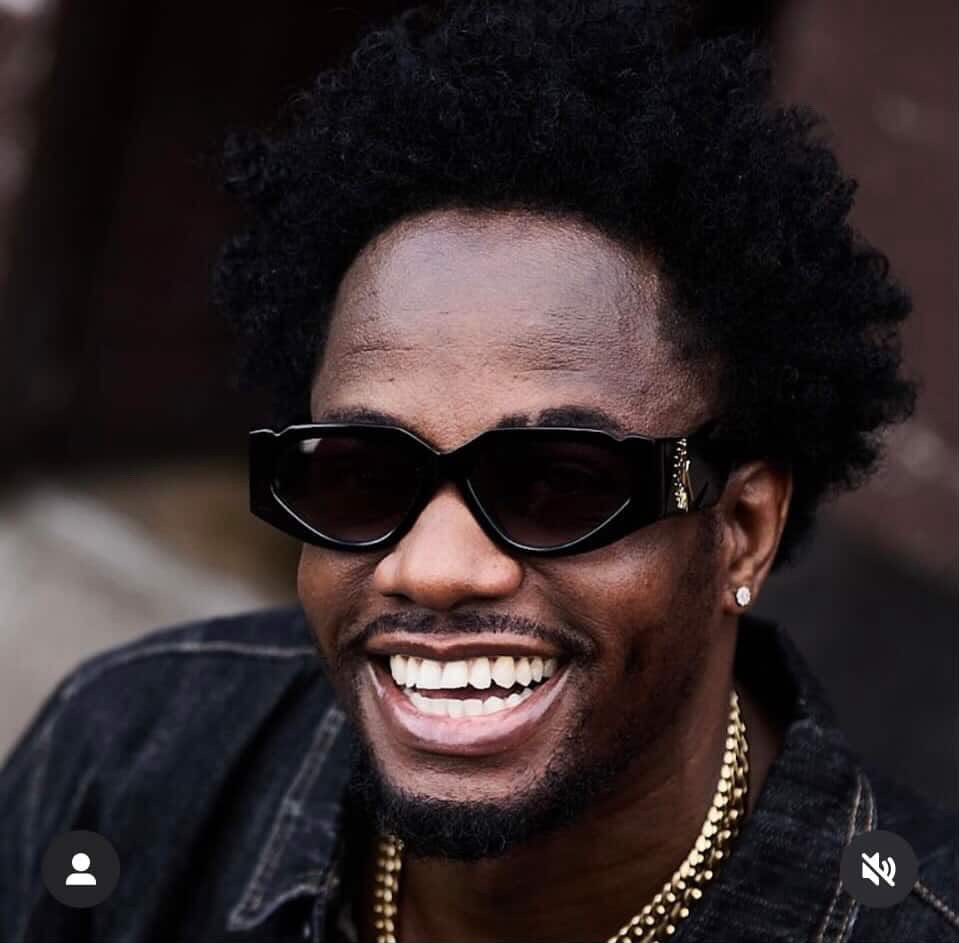 Person with curly hair wearing sunglasses, smiling, and wearing a gold chain and earrings.