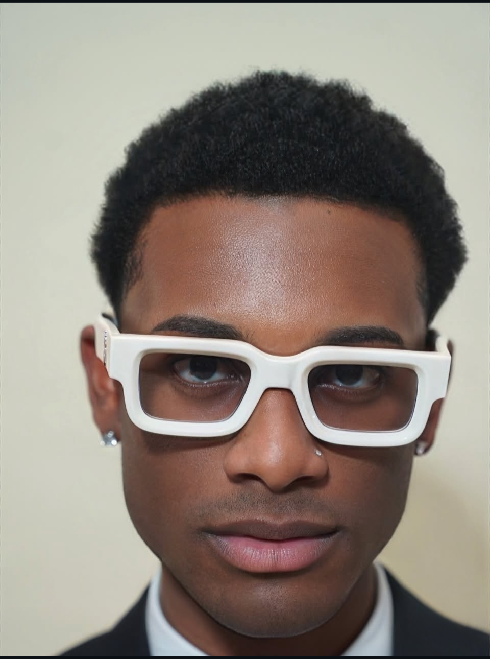 Close-up of a person wearing large white glasses, with short curly hair and earrings, facing forward.