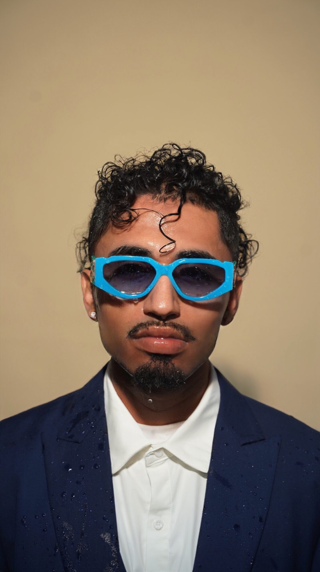 Young man with curly hair wearing blue sunglasses, white shirt, and dark blazer against a plain background.