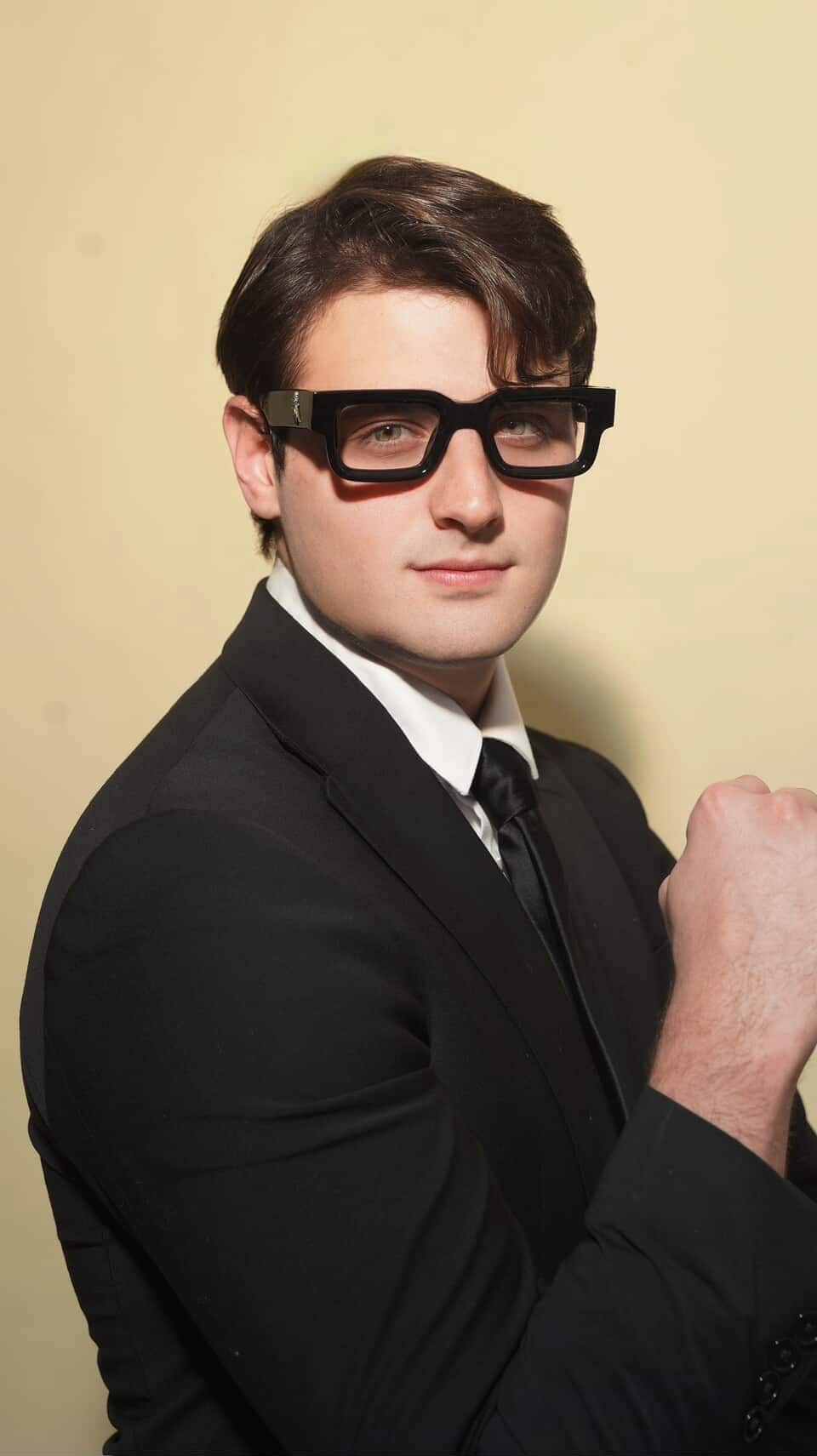 Young man in a suit and glasses, posing with a clenched fist, against a plain background.