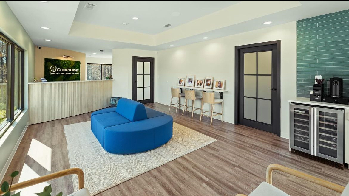Modern living room with blue sofa, white rug, wooden floor, and a wall with framed pictures, doors, and a beverage station.