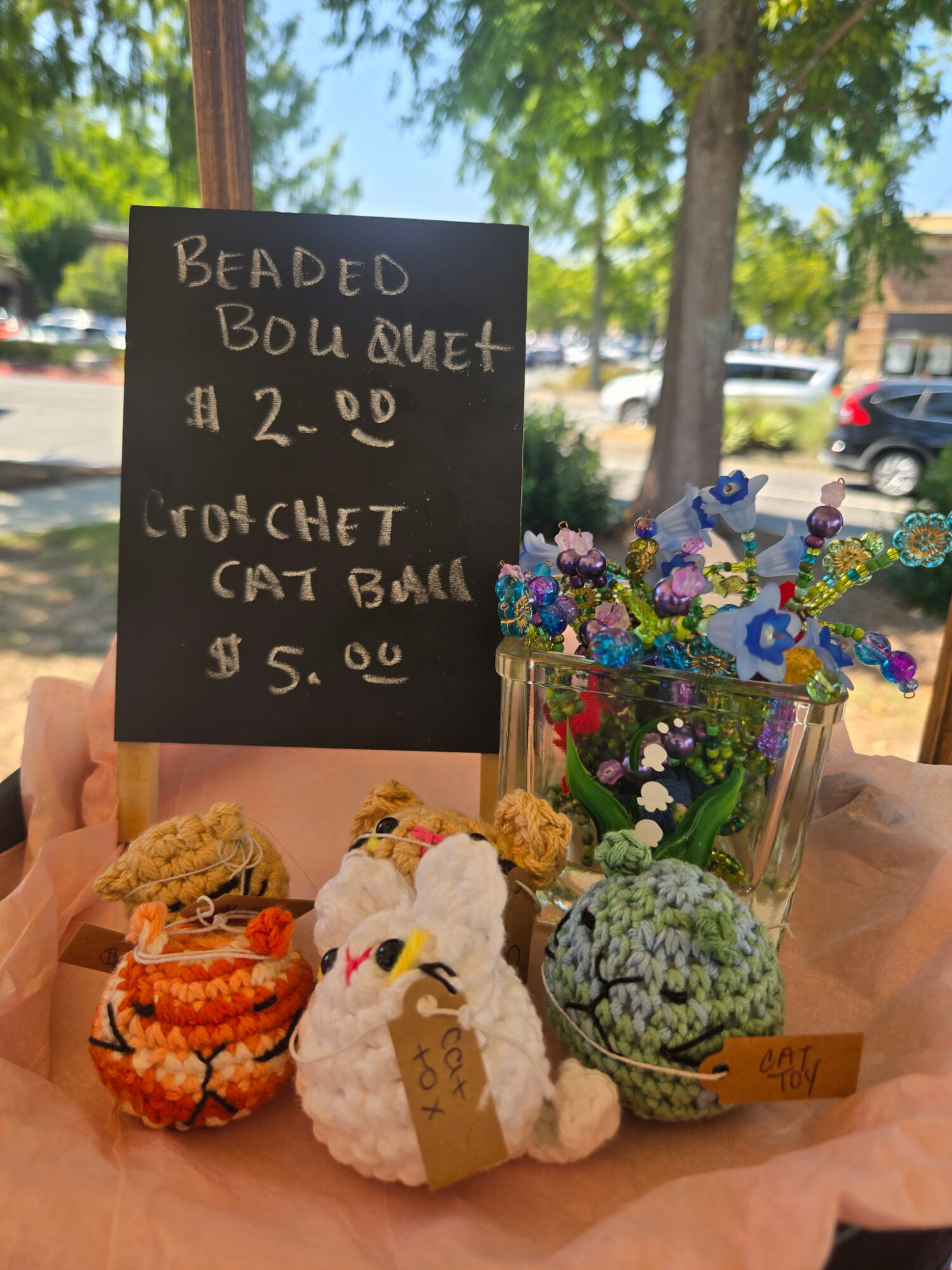 Hand holding a blackboard sign with prices, surrounded by colorful baked goods and a flower arrangement outdoors.