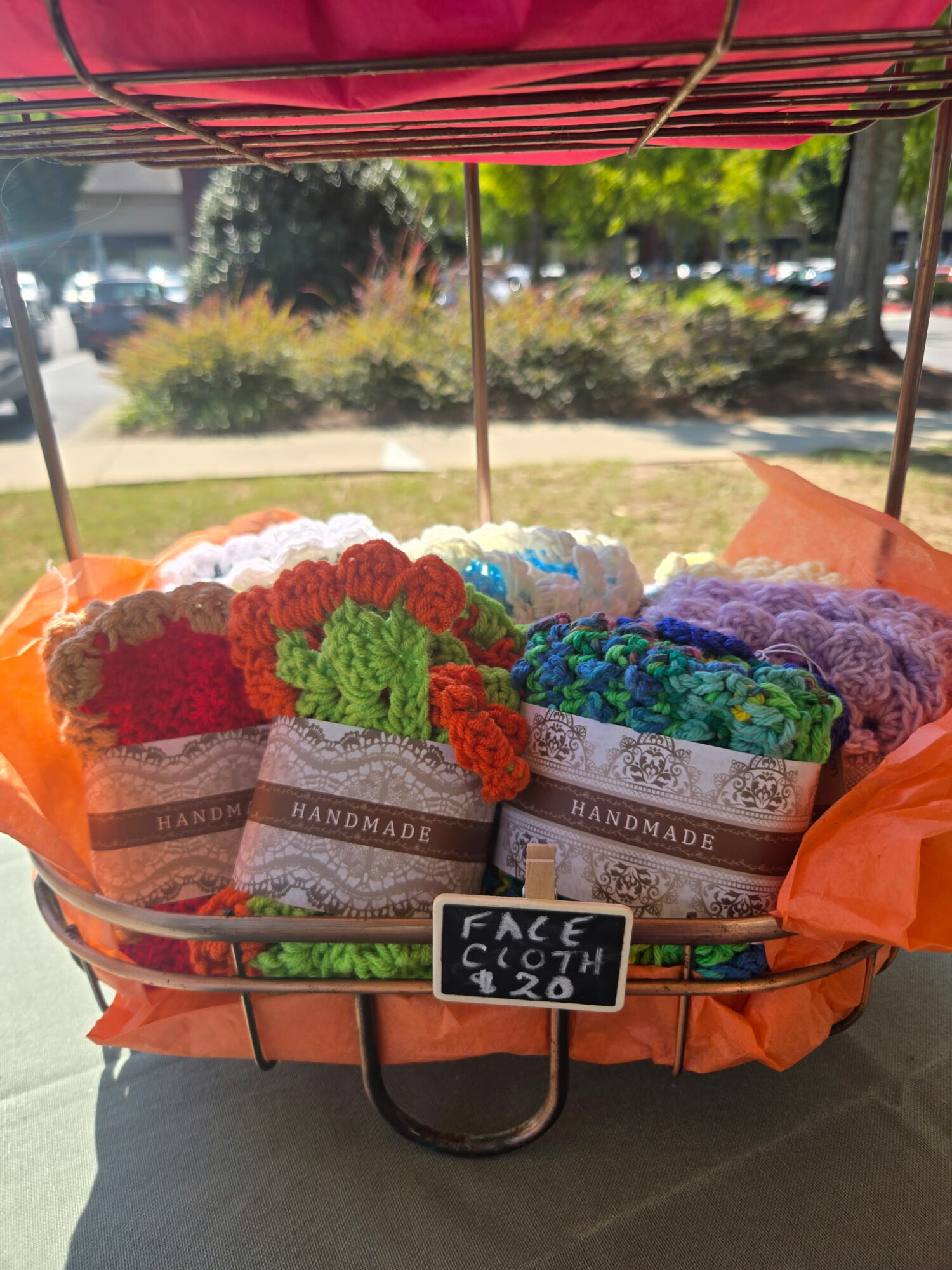 Colorful handmade knitted or crocheted items displayed in a basket outdoors, with a small blackboard sign reading 'FACE 20th'.