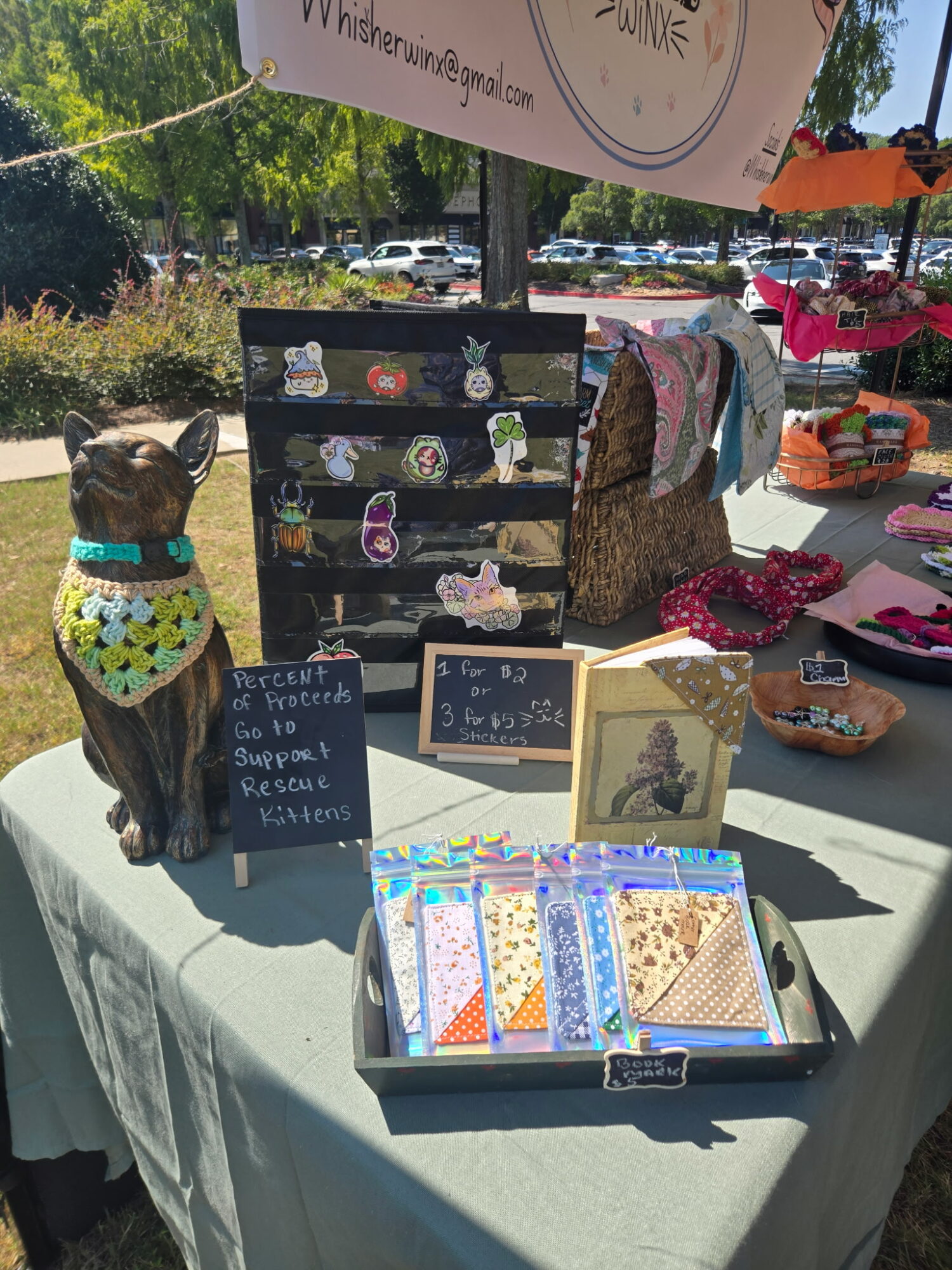 Dog wearing a colorful bandana sitting at a table with jewelry and a display stand outdoors.