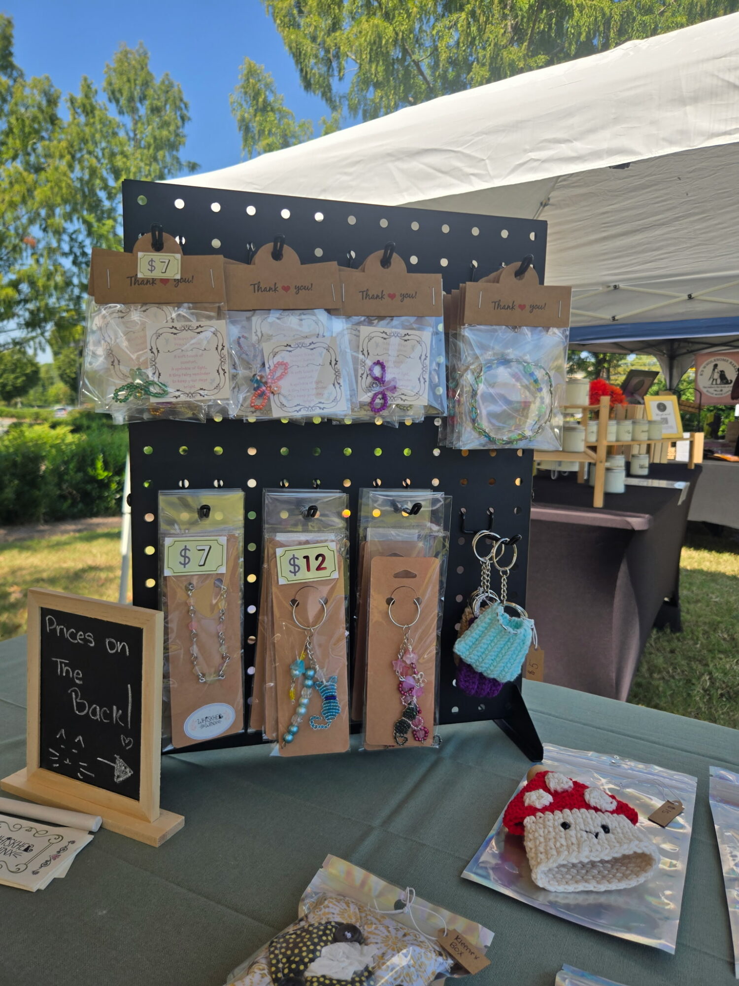 Display stand with jewelry and accessories at outdoor market, table with cake, and a chalkboard sign, under a white canopy.