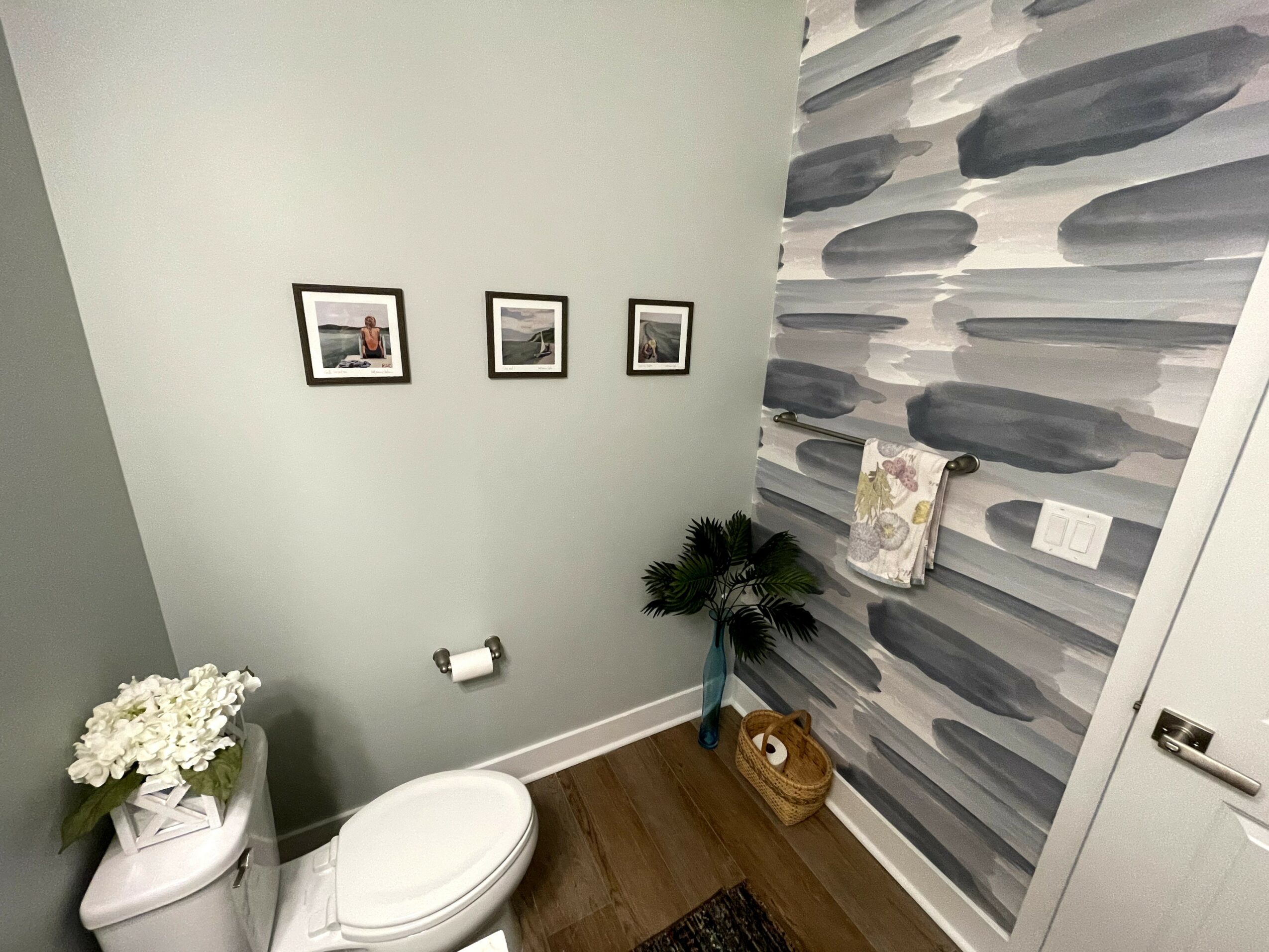 Bathroom corner with three framed pictures, a potted plant, a basket, and a towel on a patterned wall.