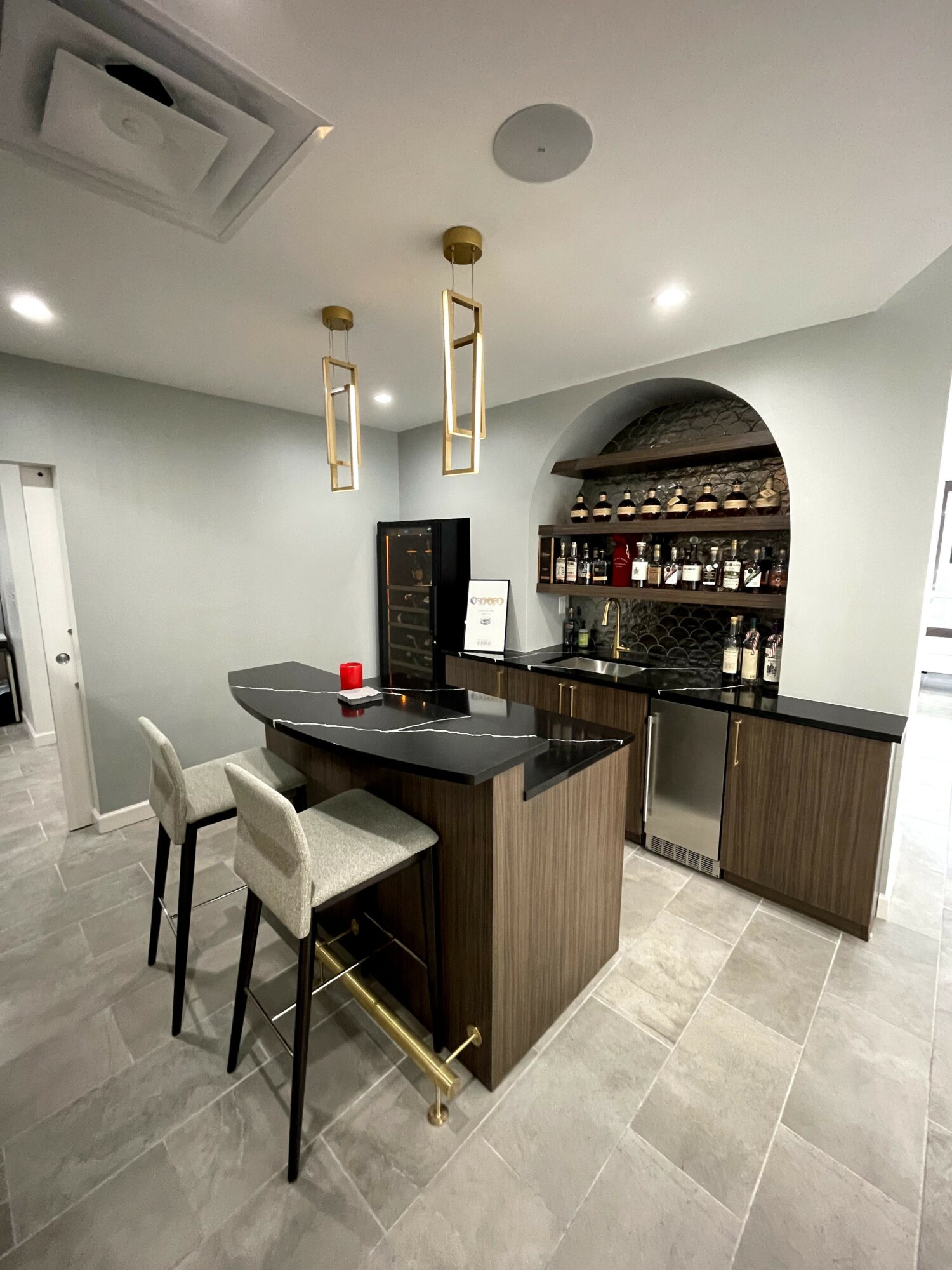 Bar area with a black countertop, two white chairs, and a wooden cabinet with shelves holding bottles and glasses.