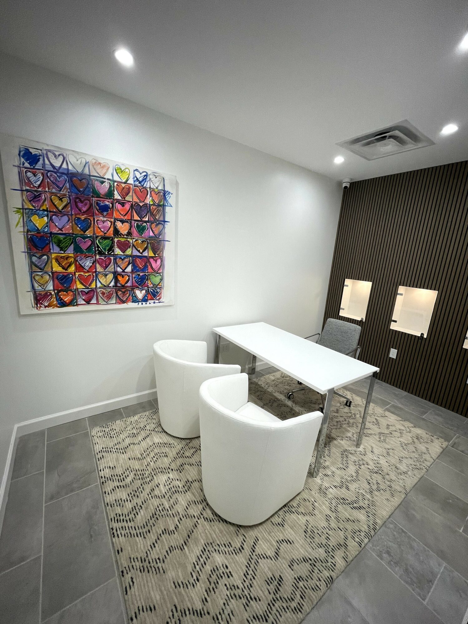 Room with white walls, colorful butterfly artwork, white table, three chairs, and a textured dark wall, with ceiling lights and air vent.