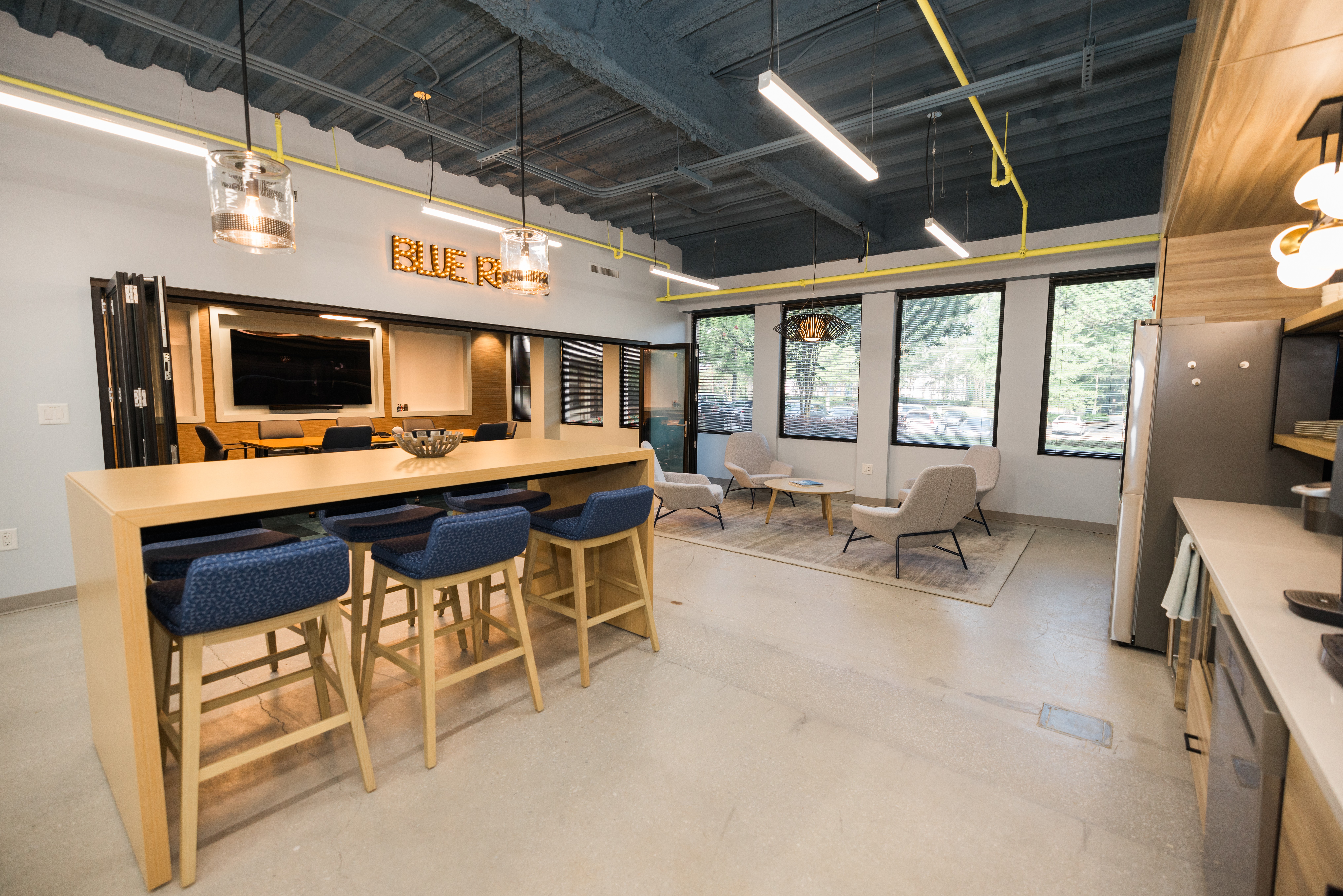 Modern lounge with bar counter, four blue chairs, seating area with armchairs, large windows, and ceiling lights.