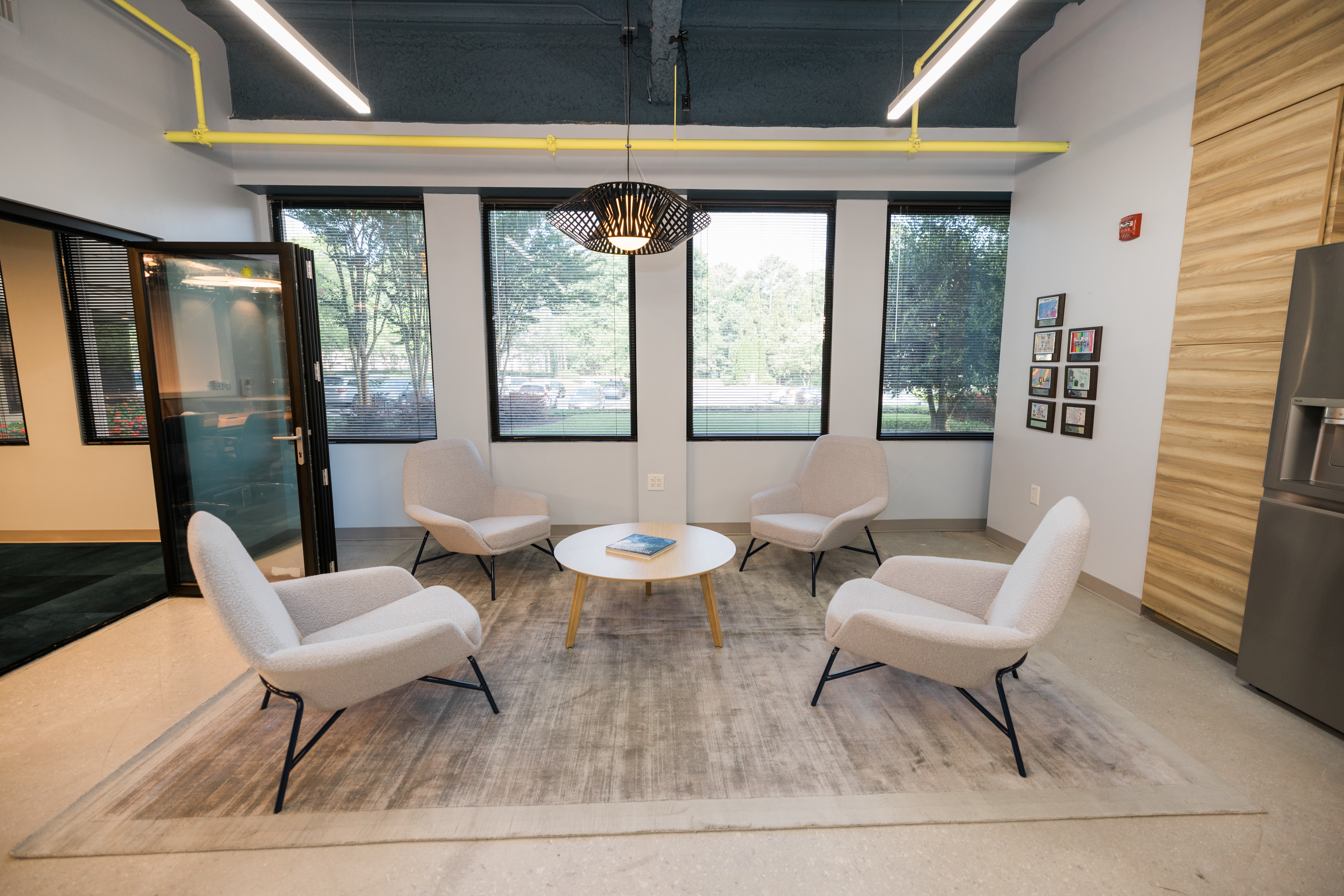 Modern lounge area with four white chairs around a round coffee table, large windows, and a gray rug.