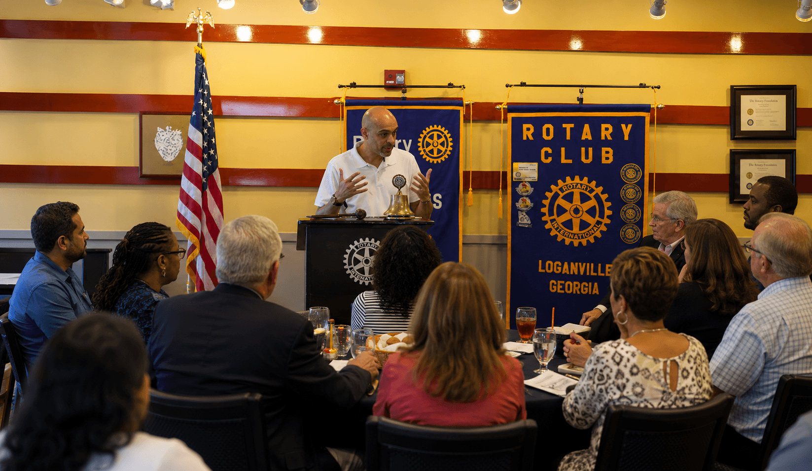 Man speaking at podium during Rotary Club meeting with seated audience, American flag, and Rotary banner in background.