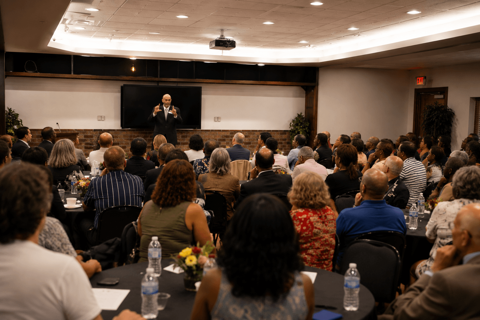 Speaker standing at podium addressing a large seated audience in a conference room, with a blackboard behind him.
