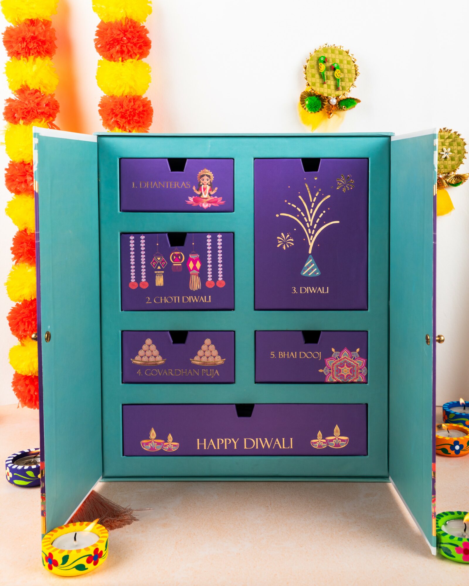 Open teal cabinet with drawers and decorations for Diwali, colorful garlands, and festive items inside and around.