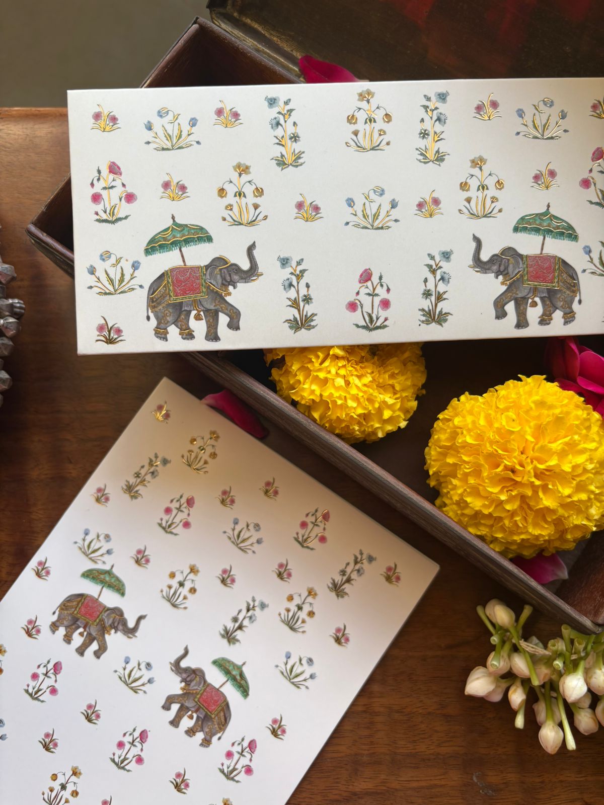 Decorative paper with elephants, umbrellas, and floral patterns, placed on a wooden surface with yellow flowers and white blossoms nearby.