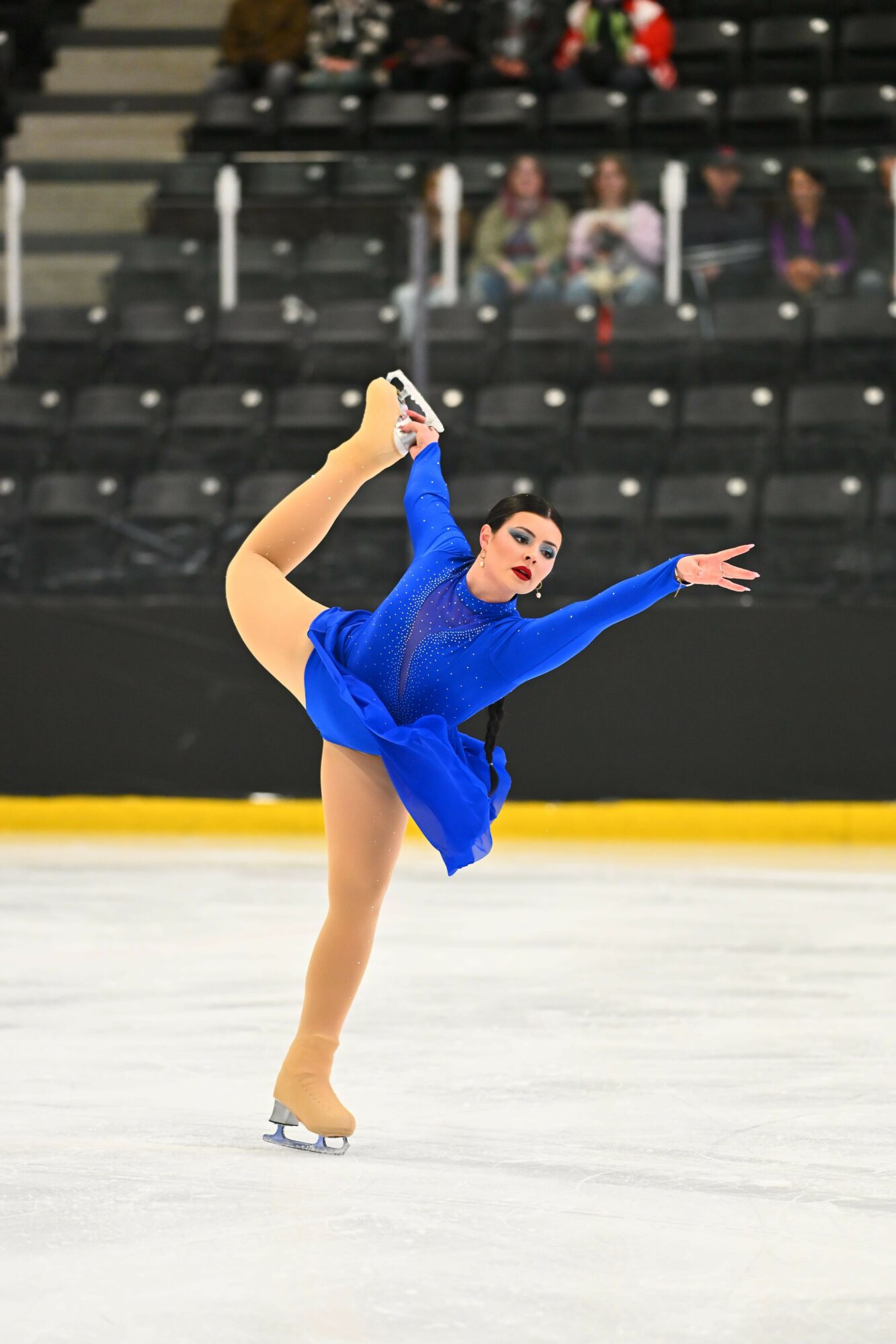 Figure skater in blue costume performing a move on ice rink with empty seats in background.