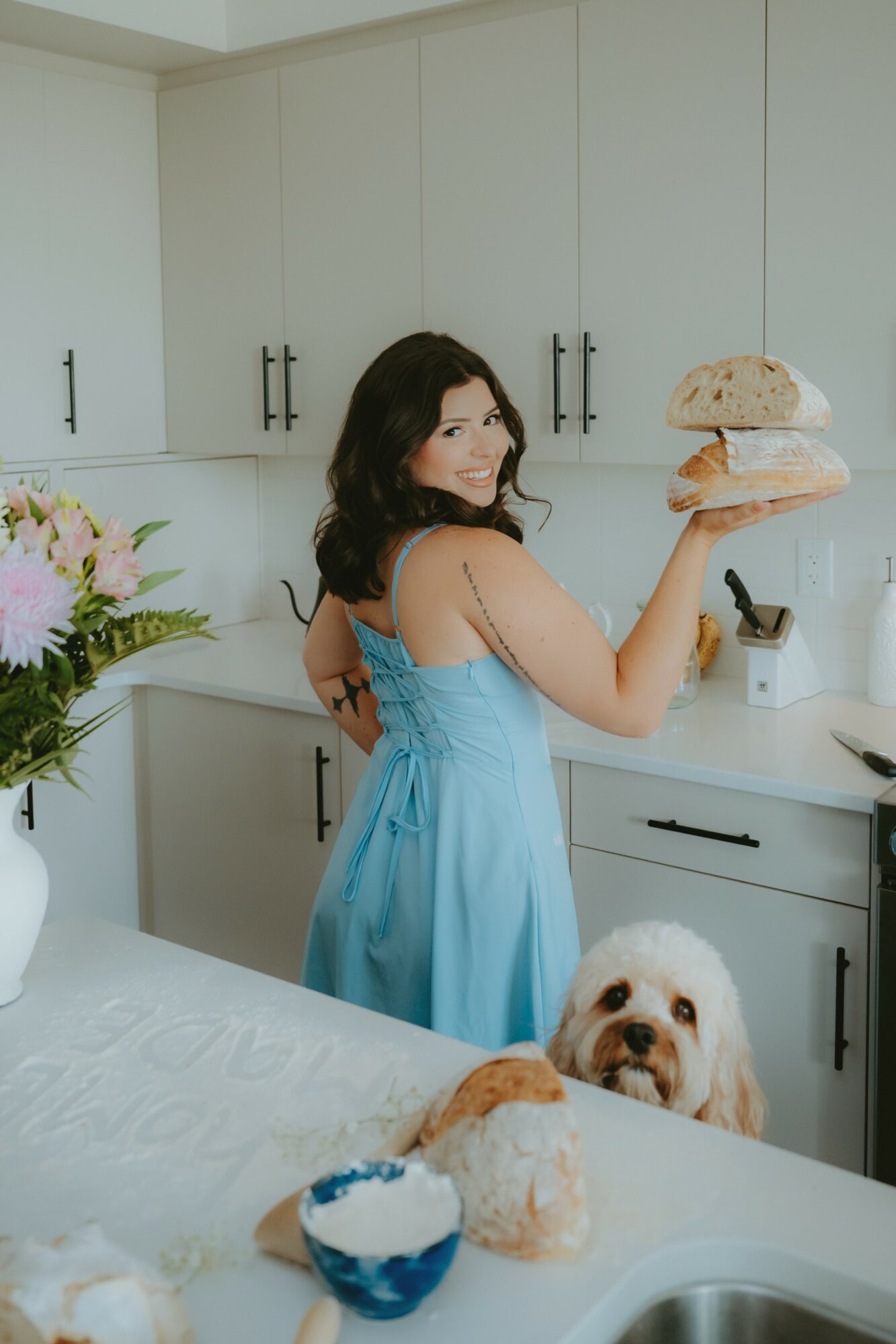 Woman in blue dress holding a sandwich in a kitchen, dog on counter, flowers in a vase, and kitchen cabinets visible.