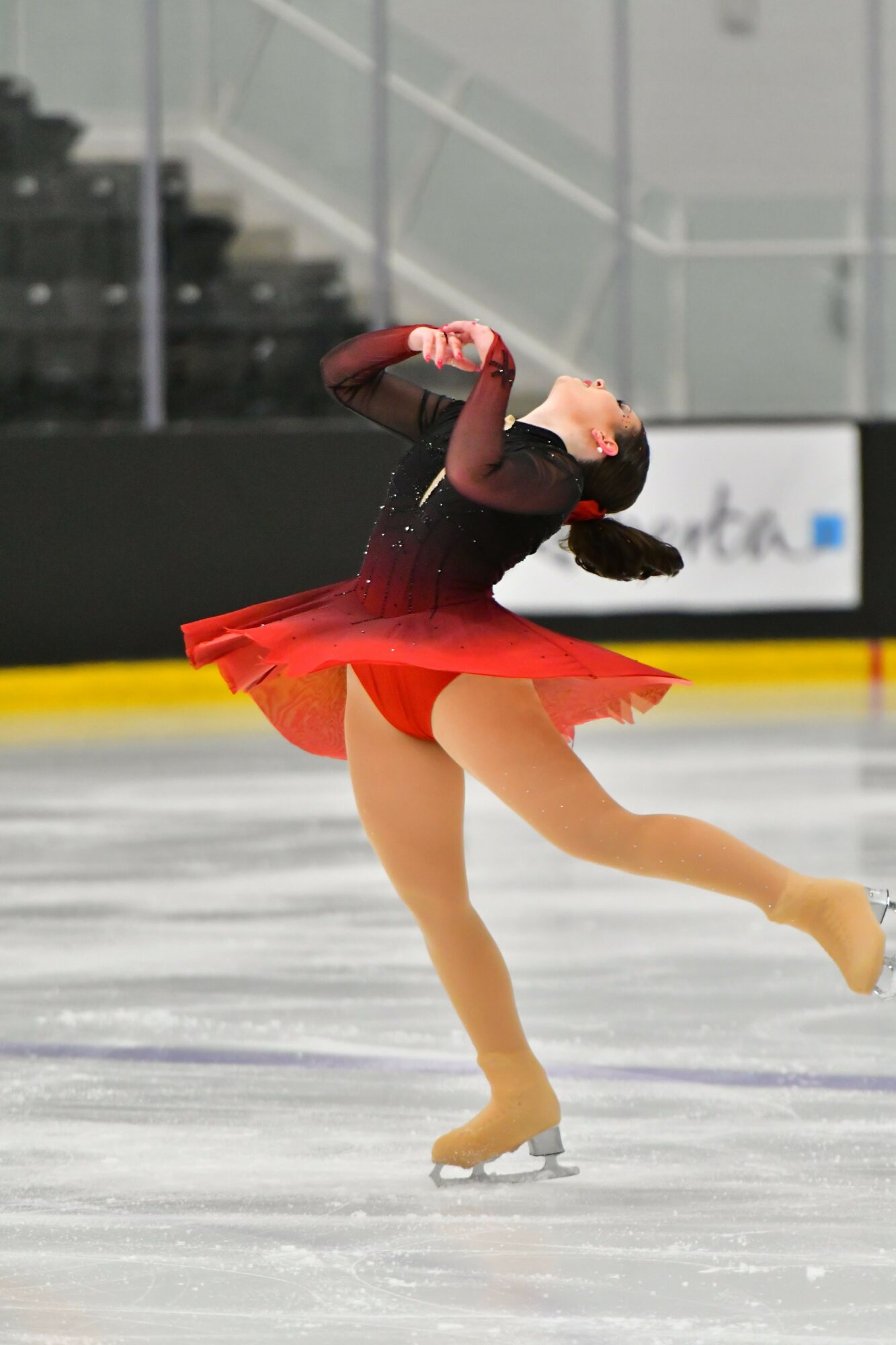 Figure skater in red dress performing a move on ice rink, with a yellow boundary and glass barrier in background.
