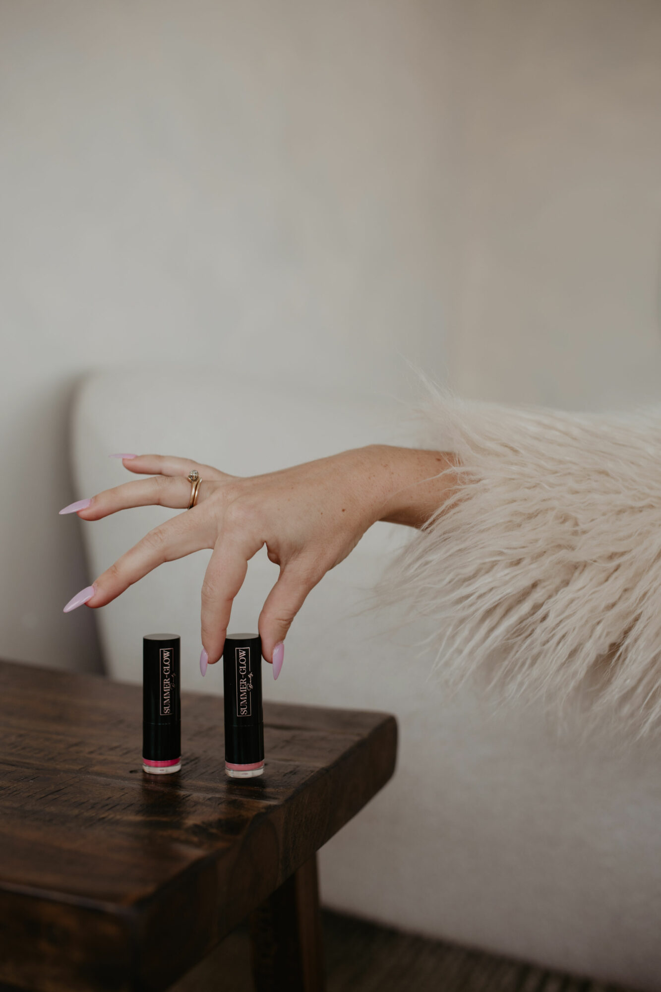 Hand with long nails reaching for two lipstick tubes on a wooden table, background is a plain wall.