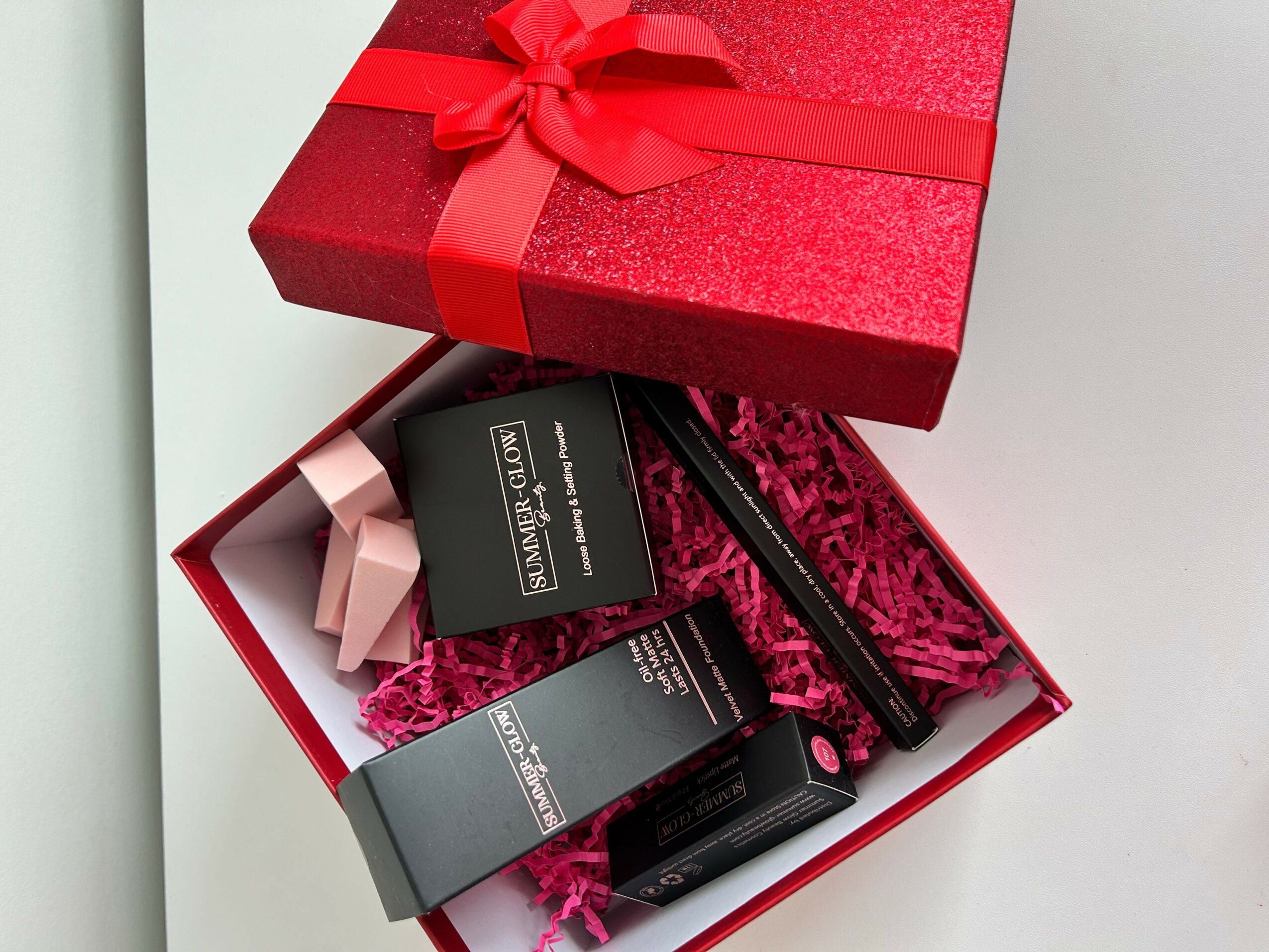 Open gift box with makeup products and pink tissue paper inside, red gift box with ribbon on top, plain white background.
