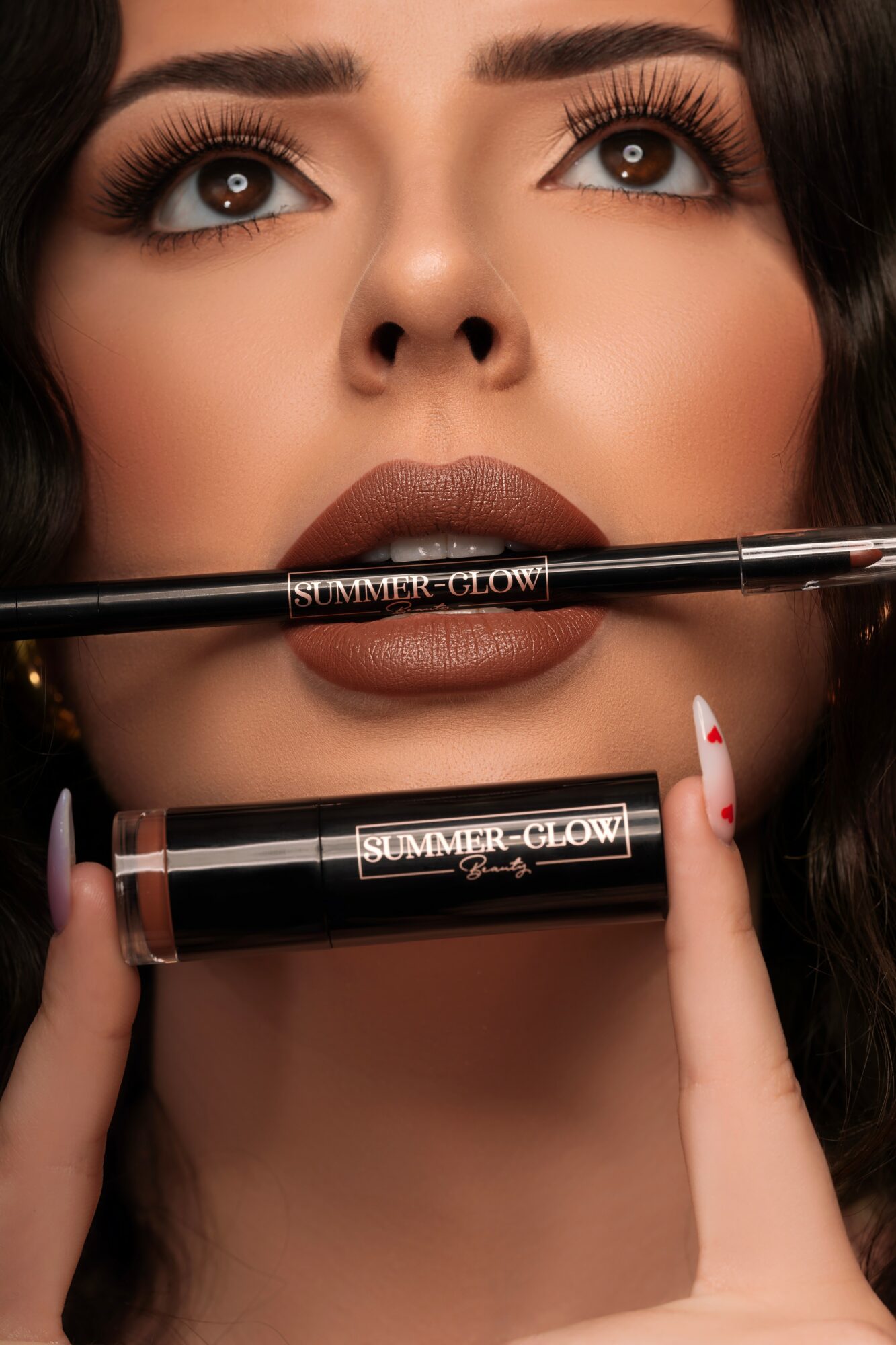 Close-up of a woman with makeup holding two lip gloss tubes near her lips, looking upward.