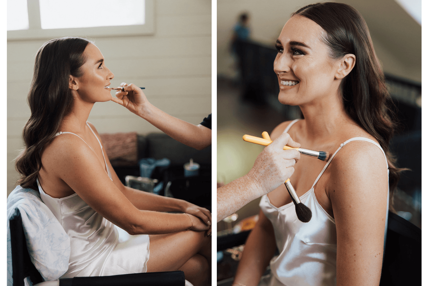 Woman getting makeup applied, smiling, sitting in a room with natural light, wearing a white satin top.