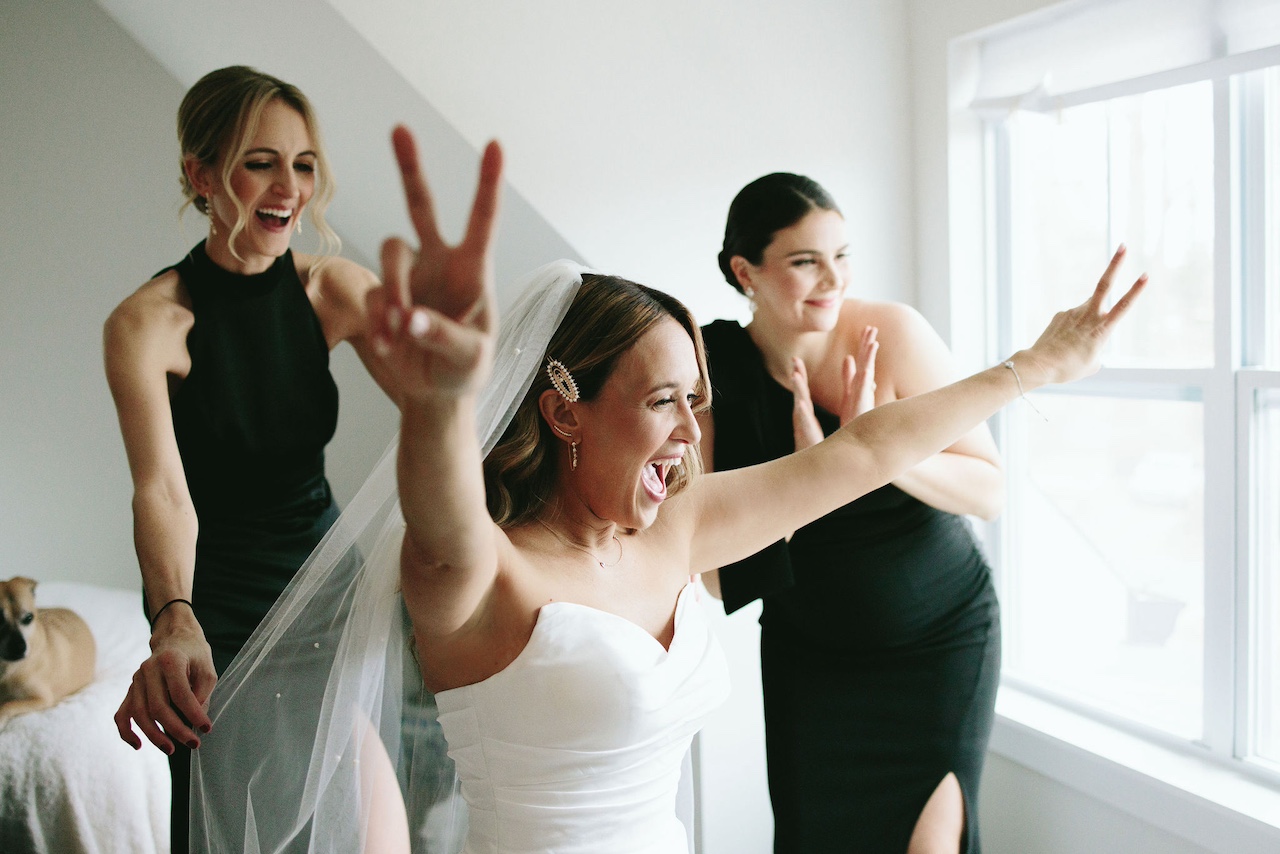 Three women, one in a wedding dress, celebrating and smiling near a window with sunlight, indoors.
