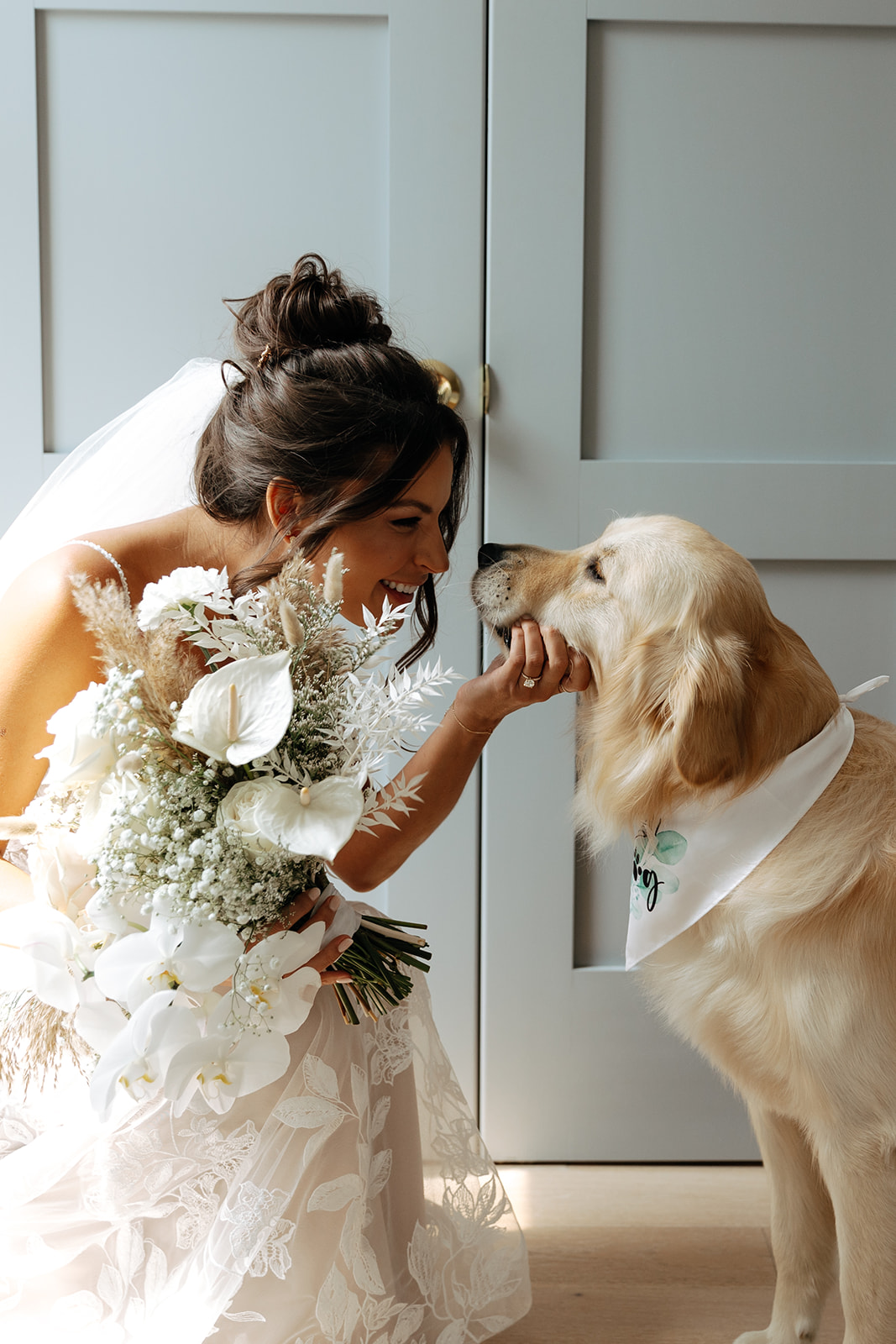Woman in wedding dress holding bouquet gently touching dog's nose, both indoors near gray cabinets.