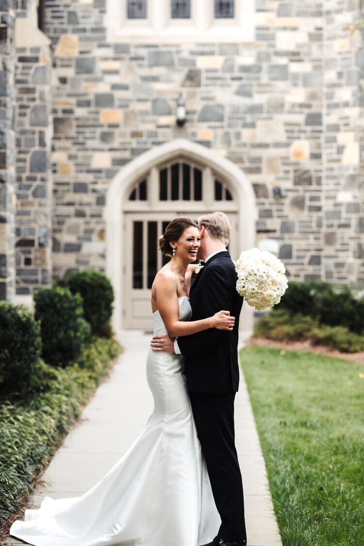 Bride and groom embrace outdoors in front of a stone building, holding a bouquet of white flowers.