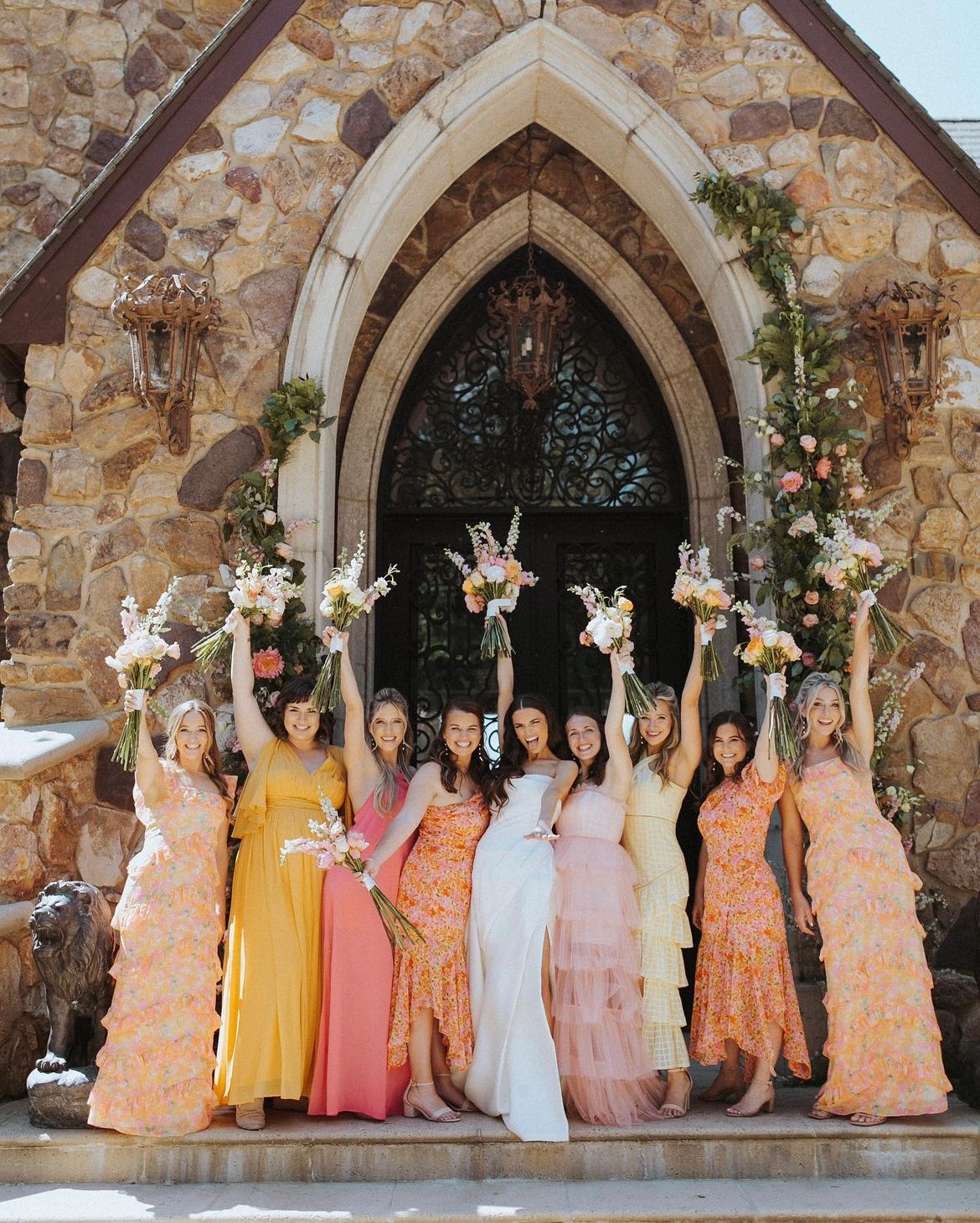 Group of women in colorful dresses standing in front of a stone church entrance, holding bouquets, celebrating a special occasion.