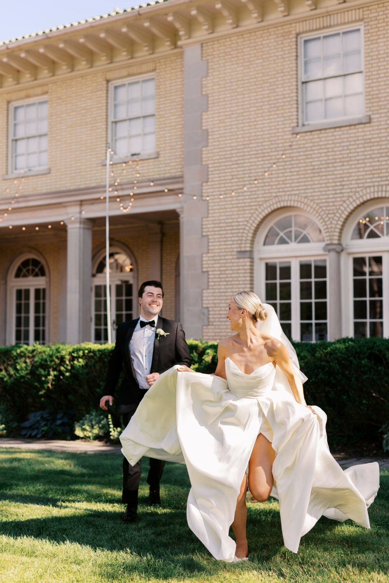 Bride and groom outdoors in front of a large brick building, bride lifting her dress, groom smiling, sunny day.