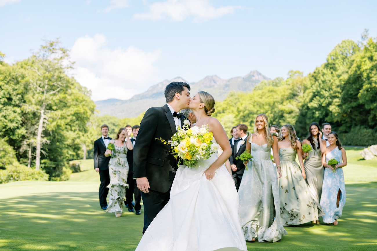 A bride and groom kiss outdoors with wedding guests in the background, trees, and mountains, sunny day.