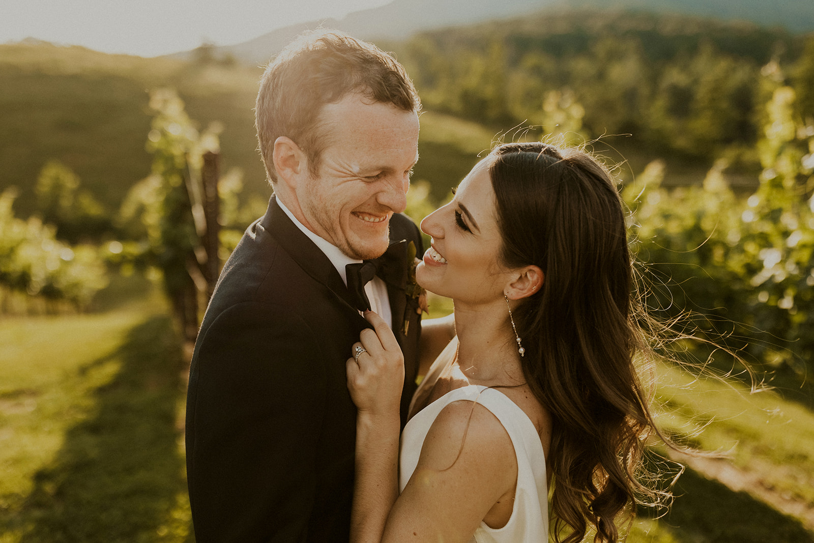 A smiling couple in formal attire outdoors with greenery and hills in the background.