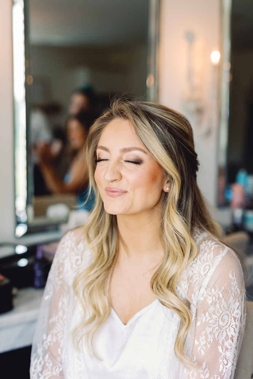 Woman with long wavy blonde hair and closed eyes, wearing a white lace top, in a mirror reflection.