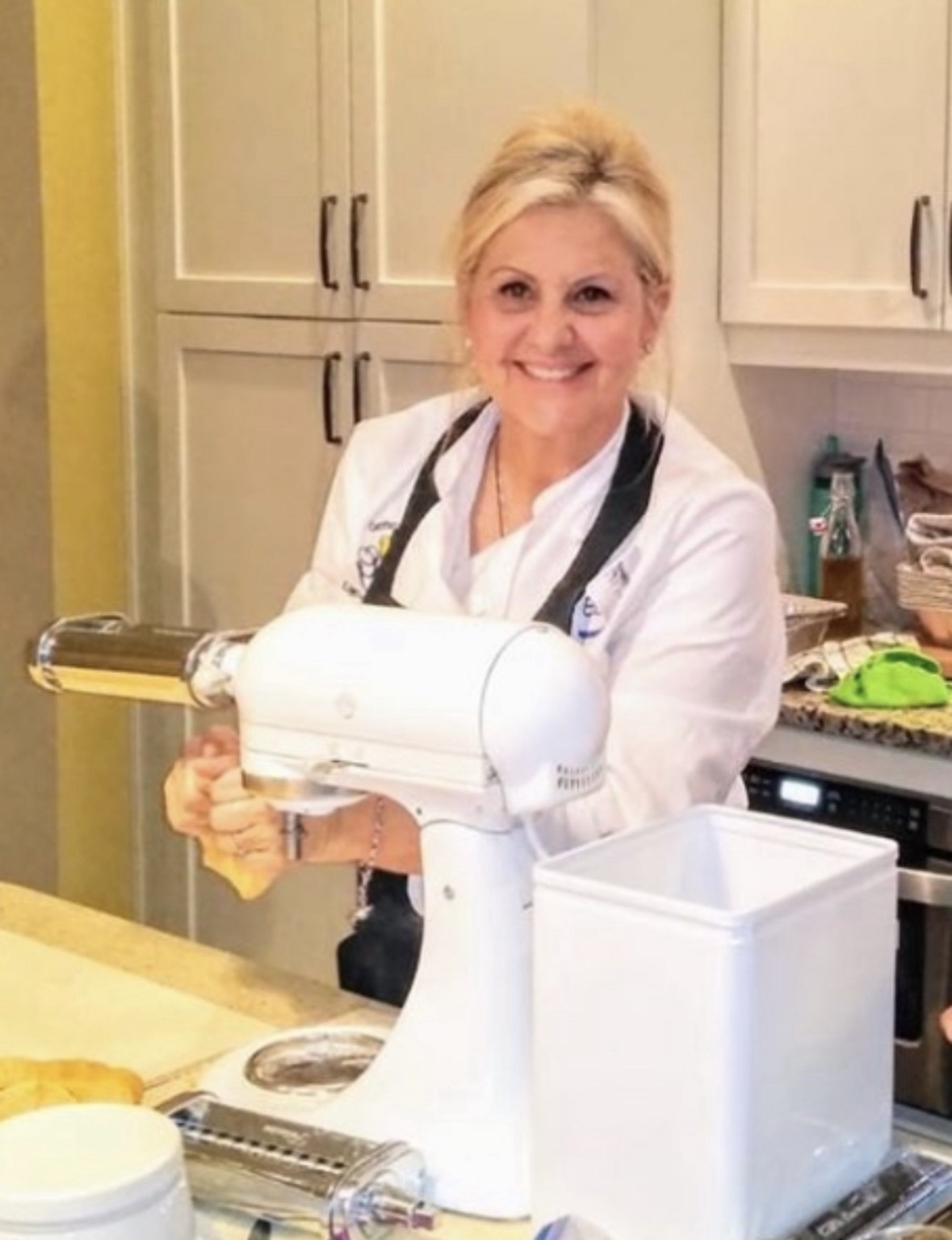 A woman in a white coat smiling in a kitchen, holding a white kitchen appliance, with cabinets and a countertop in the background.