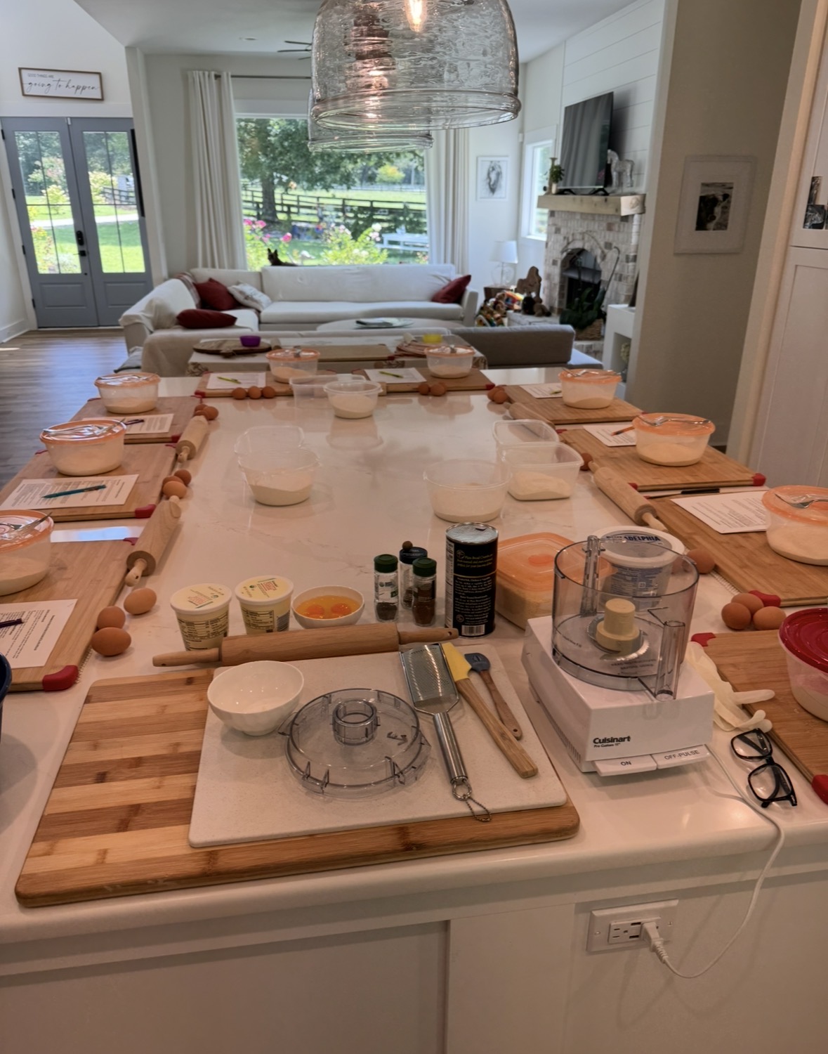 Dining table set with plates, glasses, and utensils in a bright living room with large windows and a fireplace.