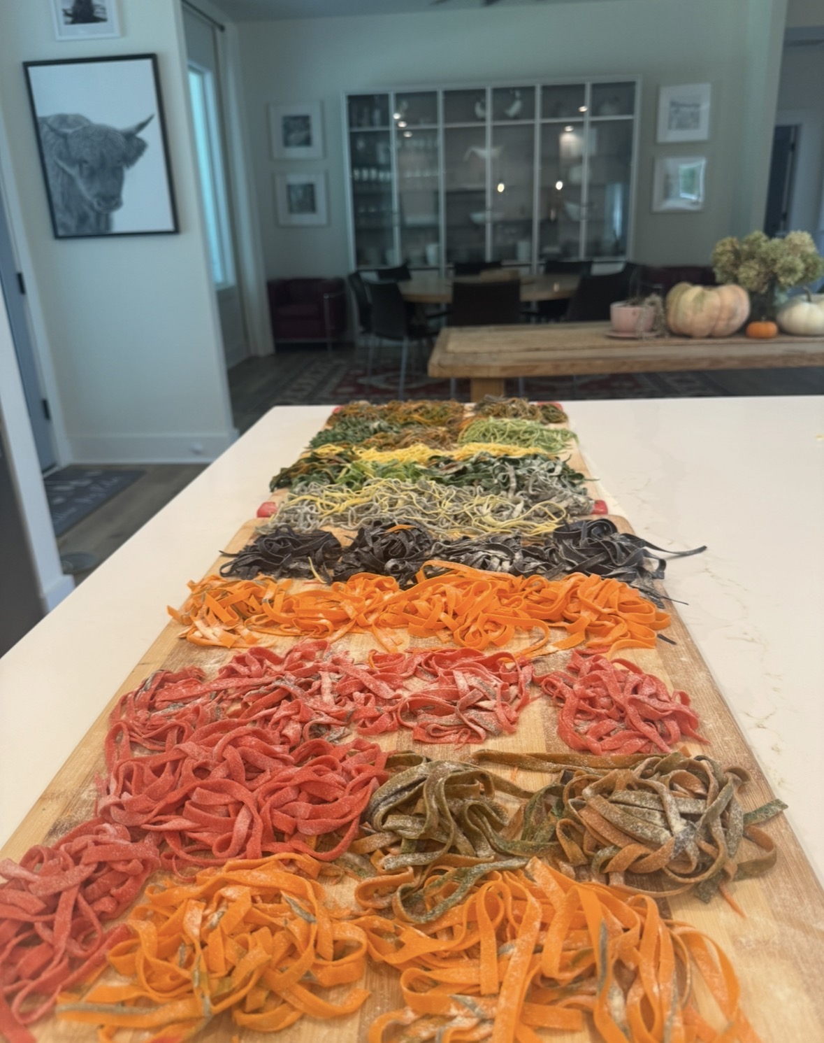 Colorful pasta arranged on a wooden surface in a kitchen, with a living room and window in the background.