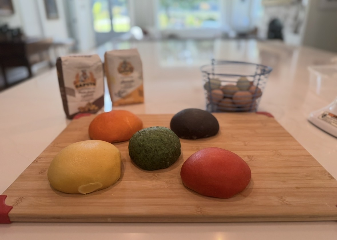 Colorful eggs and baking ingredients on a wooden cutting board in a bright kitchen, with a bowl of eggs and packages in the background.